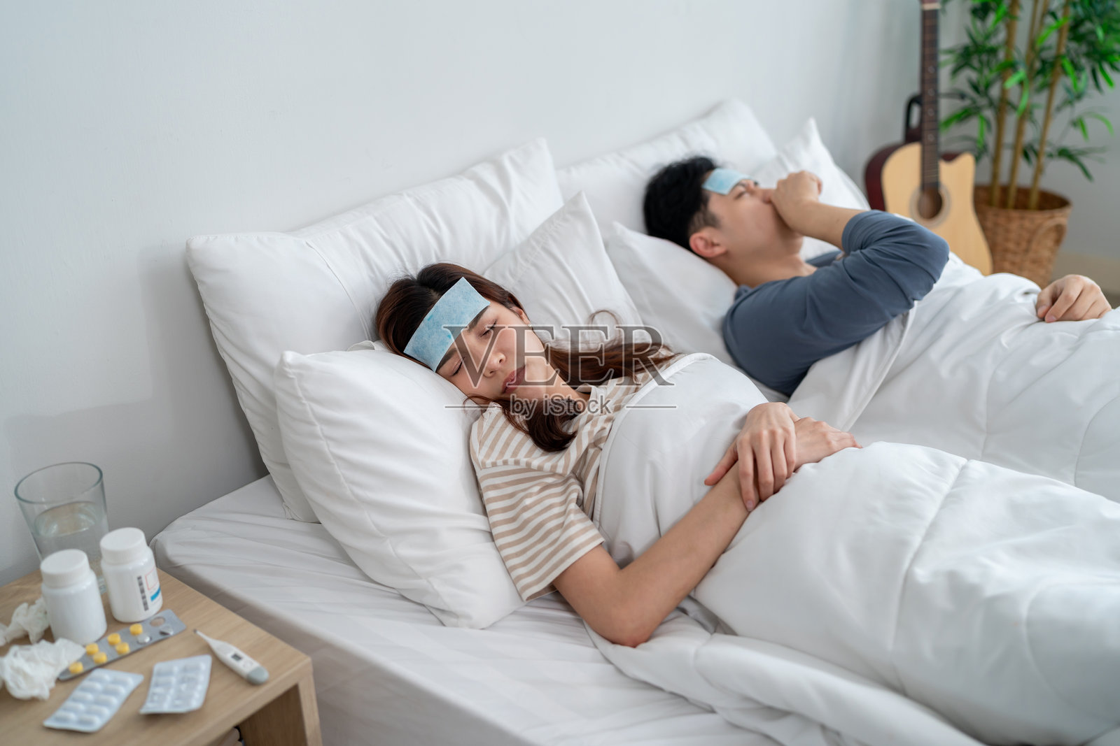 亚洲年轻情侣躺在卧室的床上，看起来生病和不适。迷人的男女额头上贴着退烧贴，早晨在睡觉时因发烧和头痛而痛苦。照片摄影图片