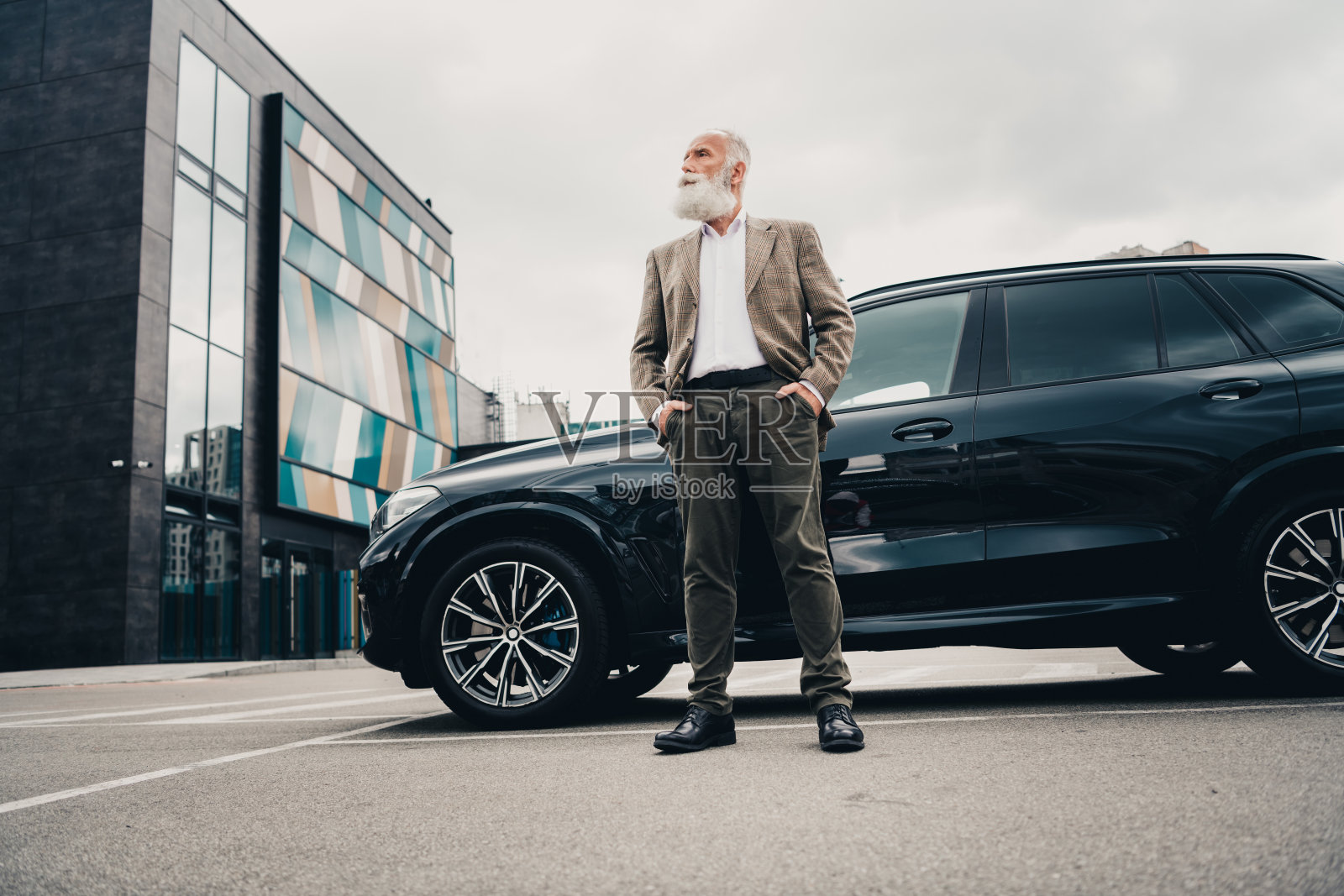 自信的成熟商人在城市环境中穿着正式服装，站在豪华车辆旁，天气阴云密布。照片摄影图片