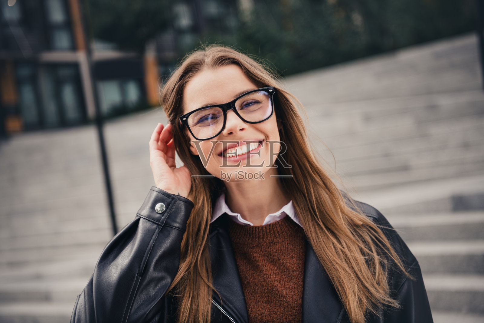 时尚的年轻女性戴着眼镜，在城市的户外阳光明媚的日子里微笑，穿着皮夹克。照片摄影图片