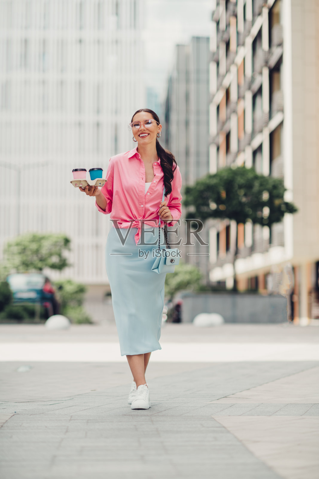 年轻的现代女性在阳光明媚的天气中，穿着时尚休闲服装，走在城市街区，手里提着咖啡杯。照片摄影图片