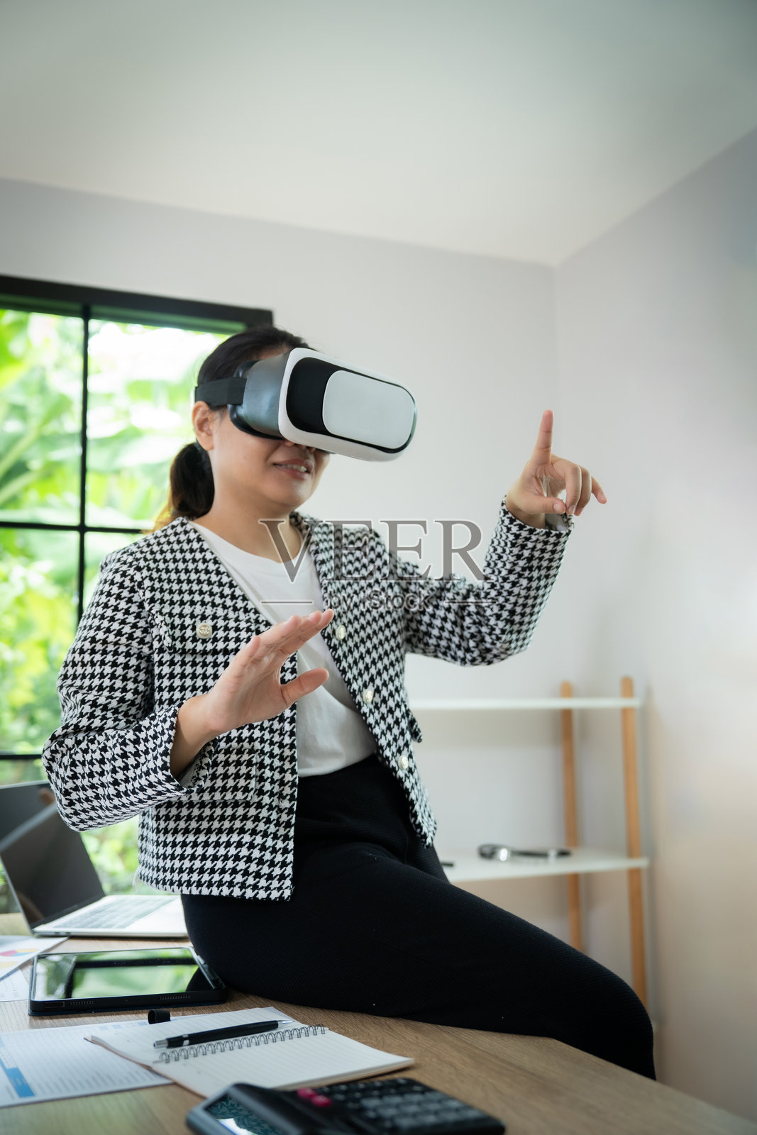 一位穿着黑白夹克的女性正在戴着虚拟现实头盔玩视频游戏。她面带微笑，向镜头挥手。照片摄影图片