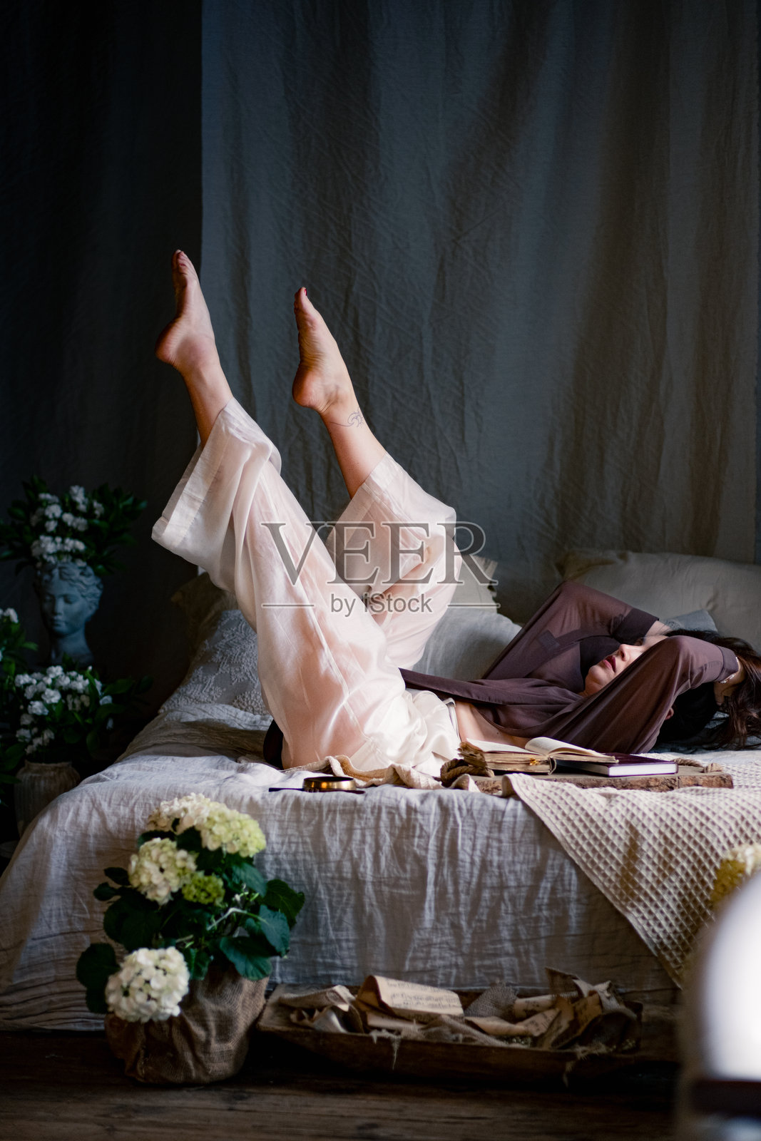 美丽的年轻女性在房间的床上摆姿势，周围有很多花。背景是复古风格的砖墙。照片摄影图片