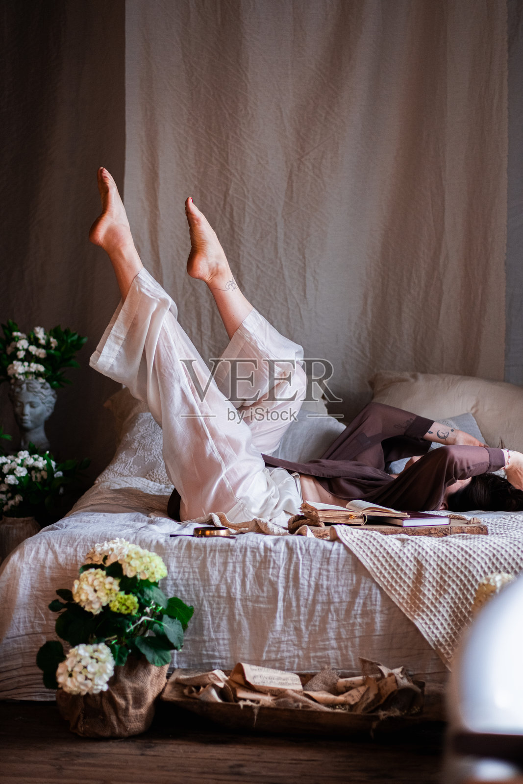 美丽的年轻女性在房间的床上摆姿势，周围有很多花。背景是复古风格的砖墙。照片摄影图片