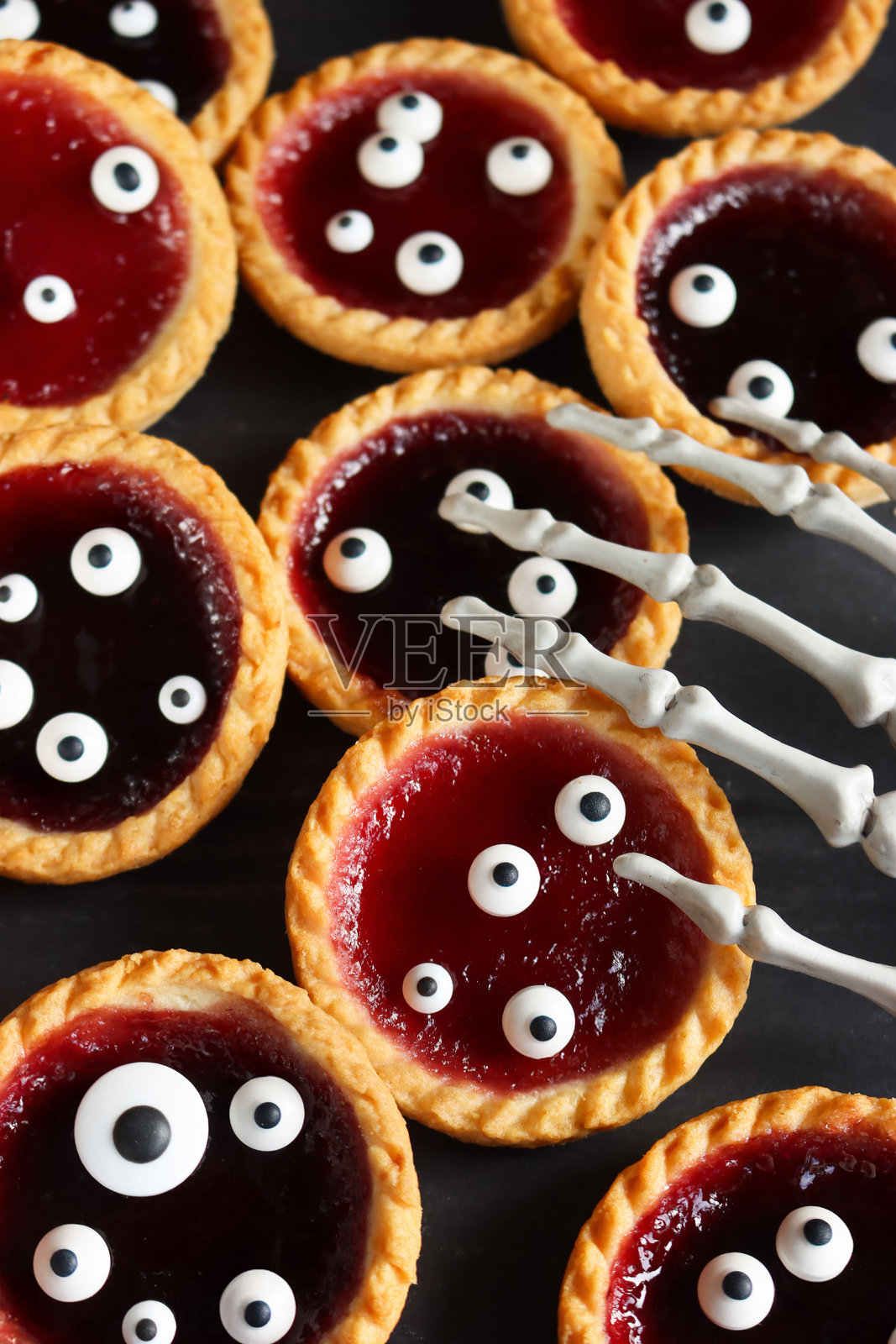 万圣节派对食物的全景图，恐怖的果酱挞，骷髅手伸向填有黑加仑和苹果或草莓果酱的酥皮派，上面装饰有翻眼糖霜，斑驳的黑色背景，俯视角度。照片摄影图片