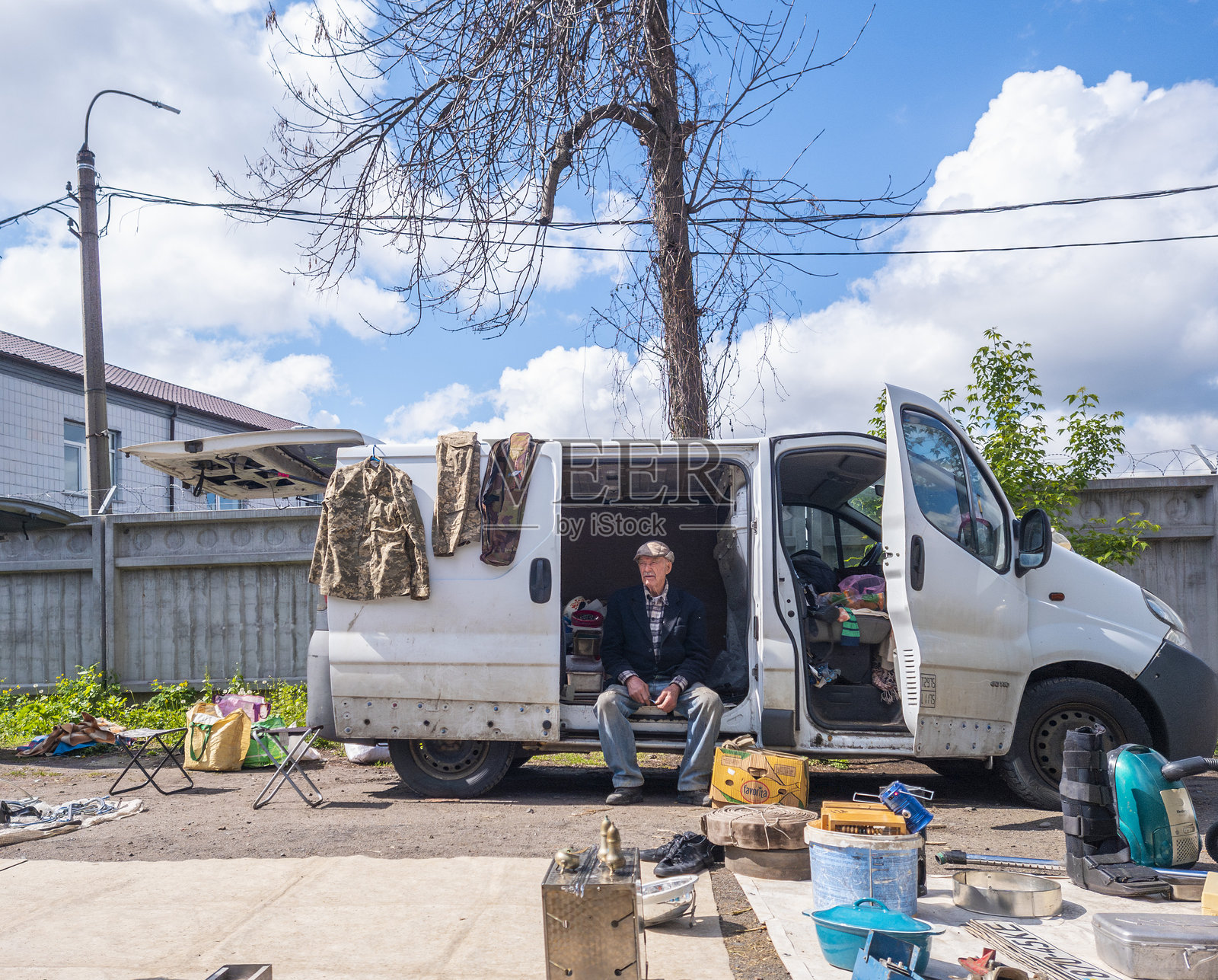 一位老年男子坐在白色面包车旁,户外街市上展示着二手物品、衣物和家居用品,蓝天上飘着几朵白云。照片摄影图片