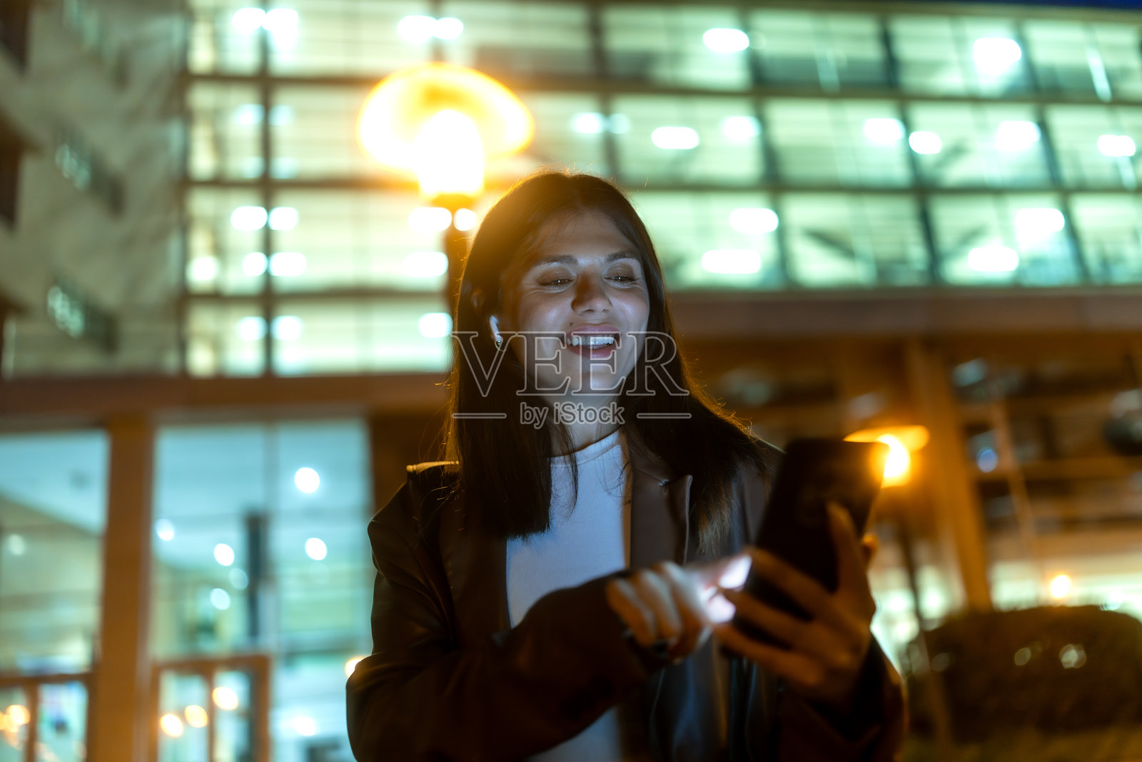 夜晚城市中，微笑的女性使用智能手机和耳机照片摄影图片