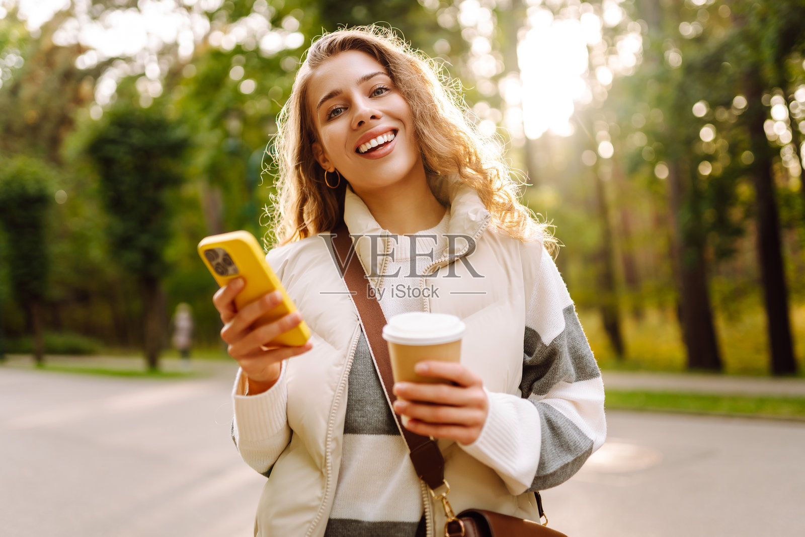 阳光明媚的公园里，手持手机的快乐女性正站在一旁，手中还拿着一杯热饮。照片摄影图片