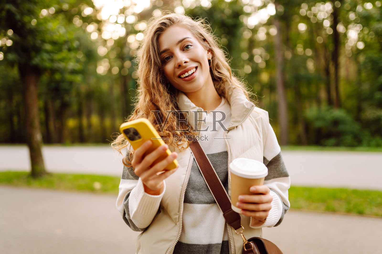 阳光明媚的公园里，手持手机的快乐女性正站在一旁，手中还拿着一杯热饮。照片摄影图片