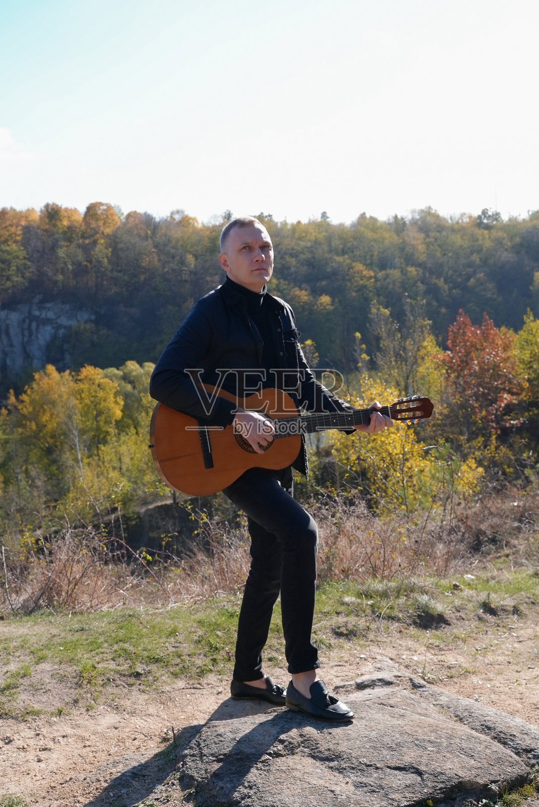 英俊的金发男子在阳光明媚的秋日户外弹奏原声吉他并放声歌唱，享受悠闲时光。这位艺术家正在学习和练习演奏乐器，周围是秋天的落叶。照片摄影图片