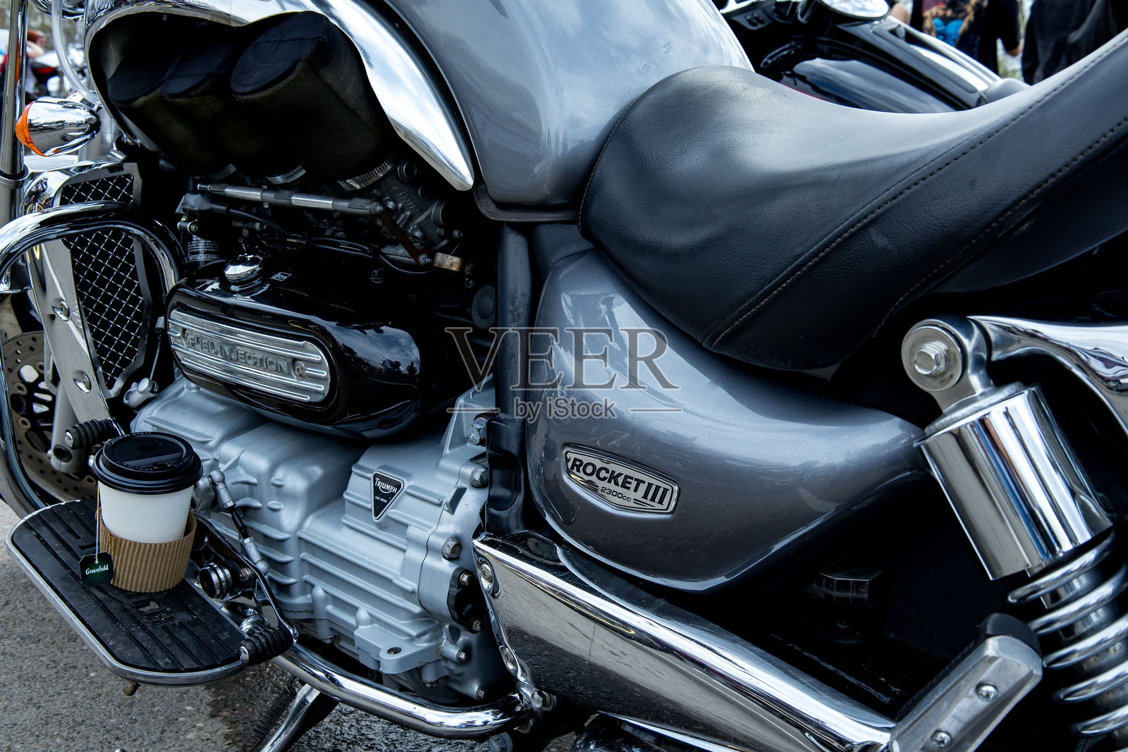 下午的阳光下，摩托车聚会中，特写了凯旋火箭三号摩托车的发动机，其脚踏板上放着一个咖啡杯。照片摄影图片