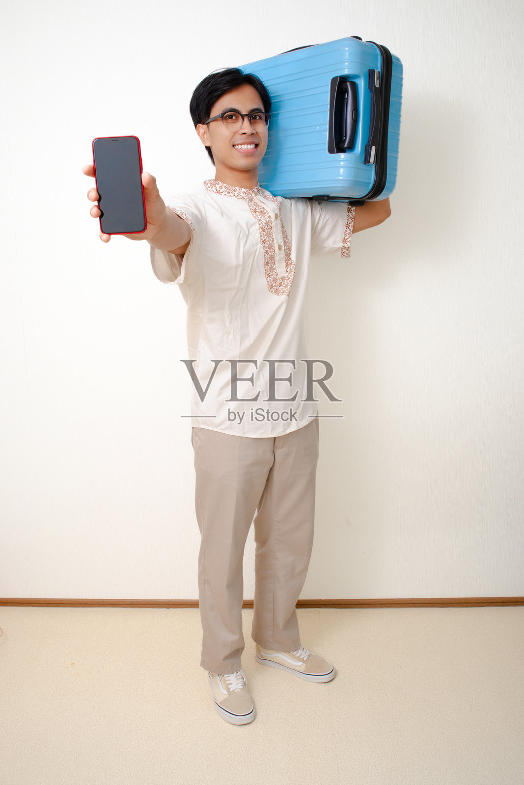 一个微笑着的亚洲穆斯林男子的全身肖像，他拉着行李箱，手里拿着一个空白的智能手机屏幕，自信地站着，表情自然友好。照片摄影图片