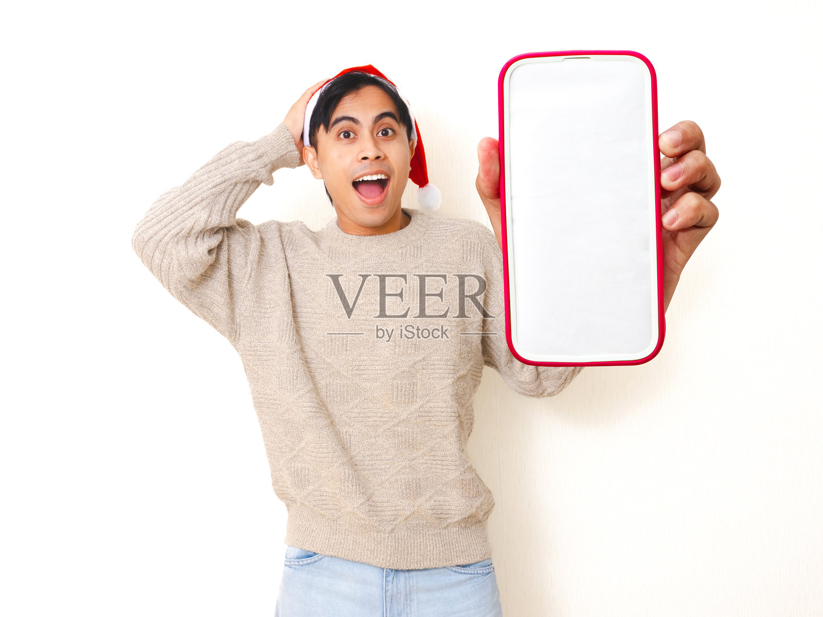 身着圣诞帽的年轻亚洲男子，在白色孤立的背景上兴奋地展示空白的智能手机模型屏幕，用于促销或广告。照片摄影图片