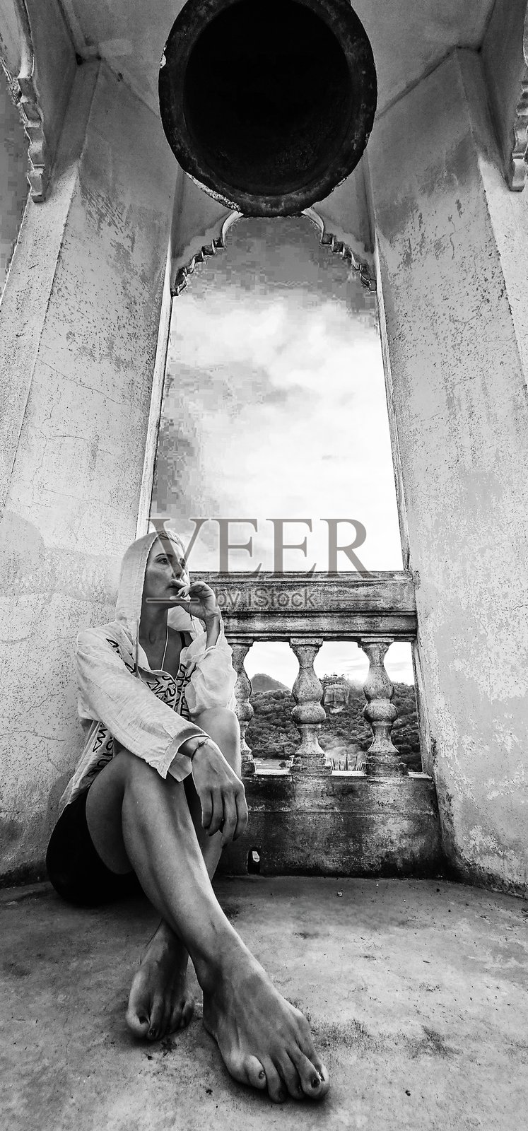 赤脚的女子身穿白色连帽衫，若有所思地坐在一块石头上。一个巨大的钟形结构和一个拱形窗户构成了云山背景。照片摄影图片