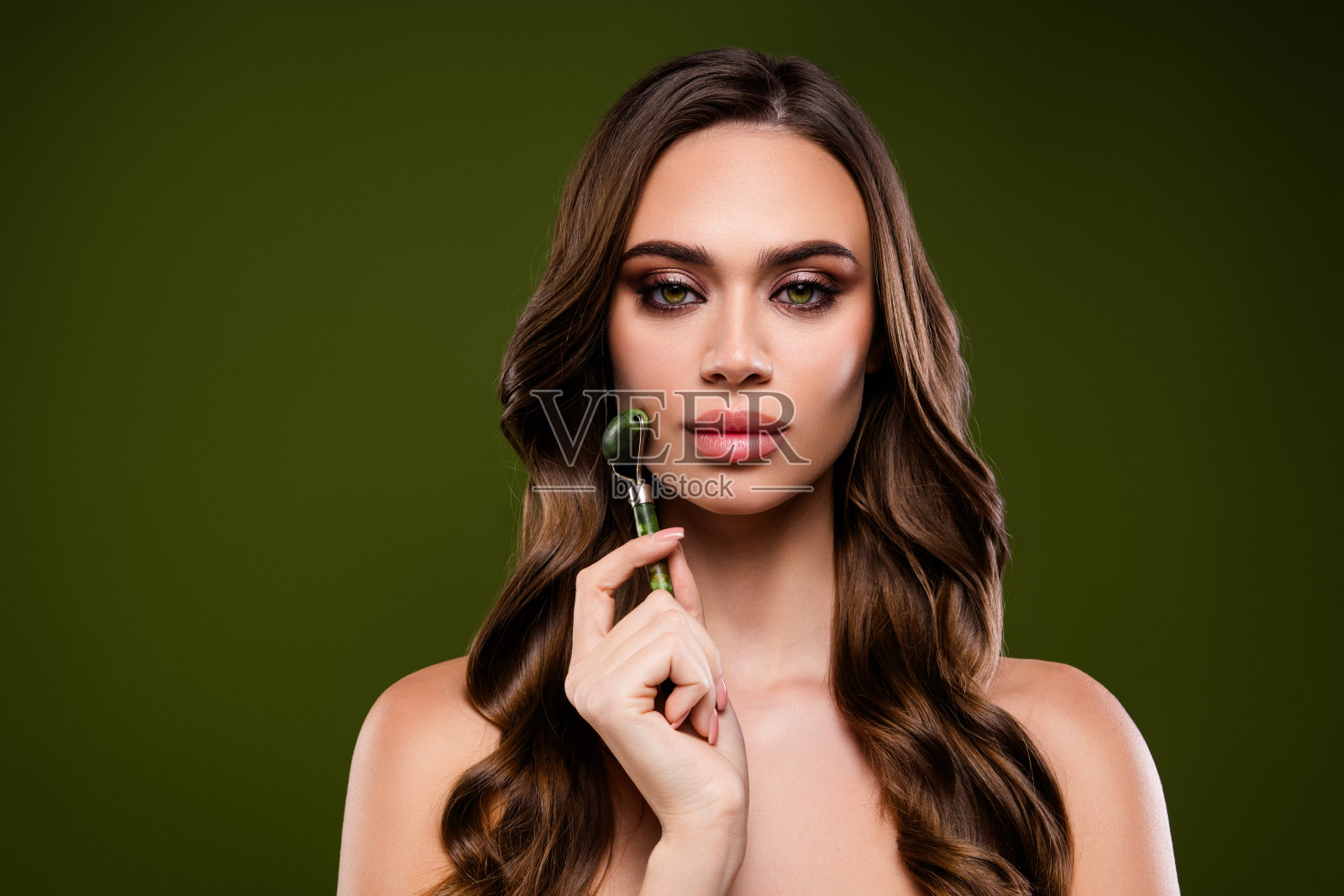 工作室里，一位有着一头棕色长发和容光焕发肌肤的美丽年轻女子正拿着绿色面部滚轮。照片摄影图片