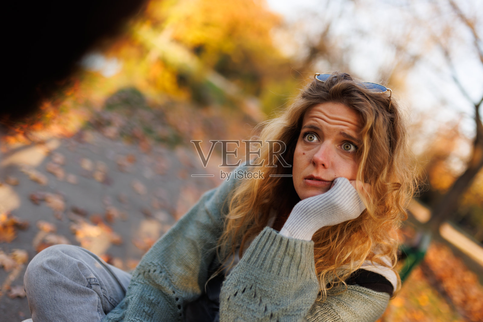 身穿舒适毛衣的女子坐在小径上，周围是秋叶，在风景如画的公园里若有所思地望着远方。照片摄影图片