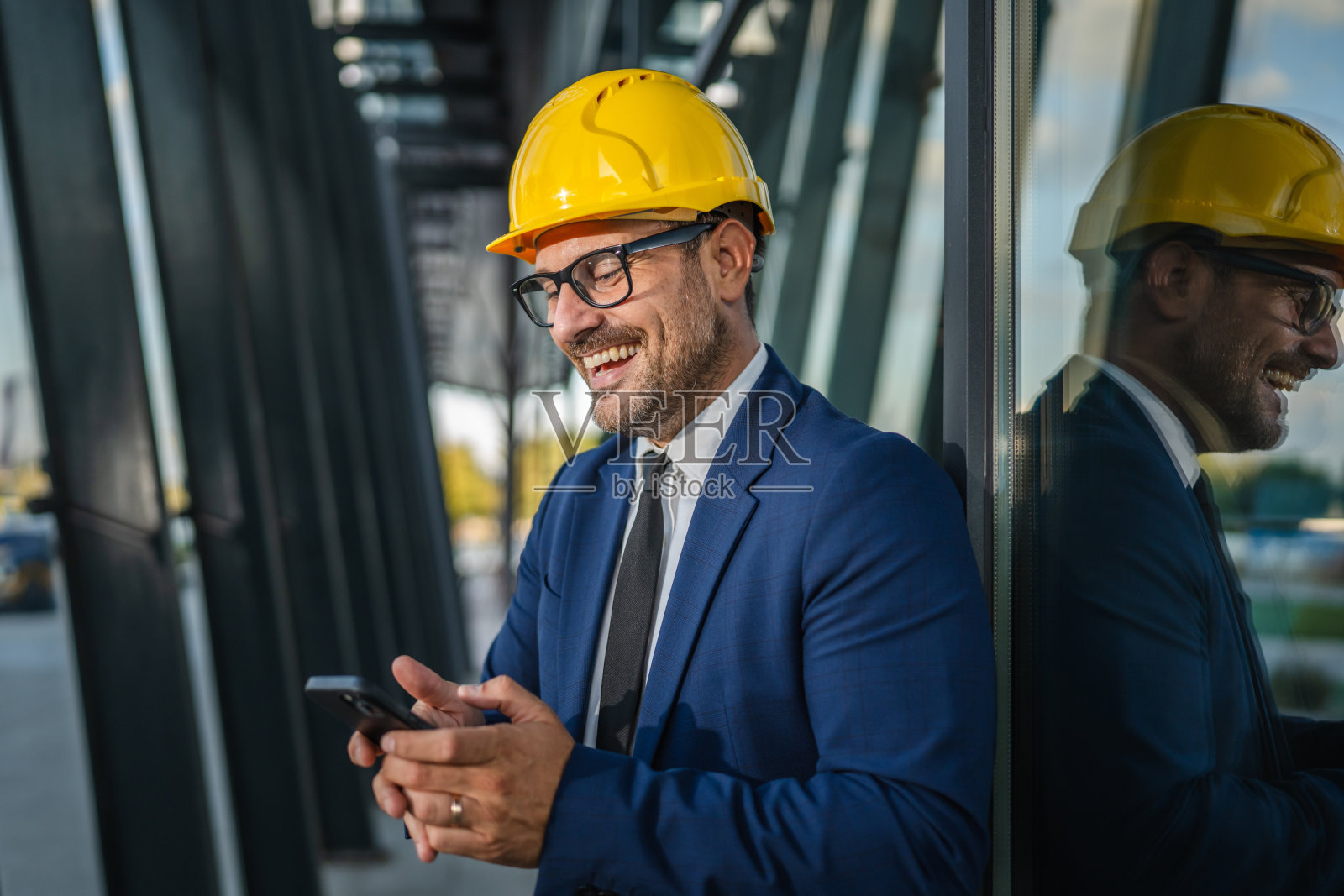 身穿西装、头戴安全帽的建筑师在现代建筑前使用智能手机。照片摄影图片