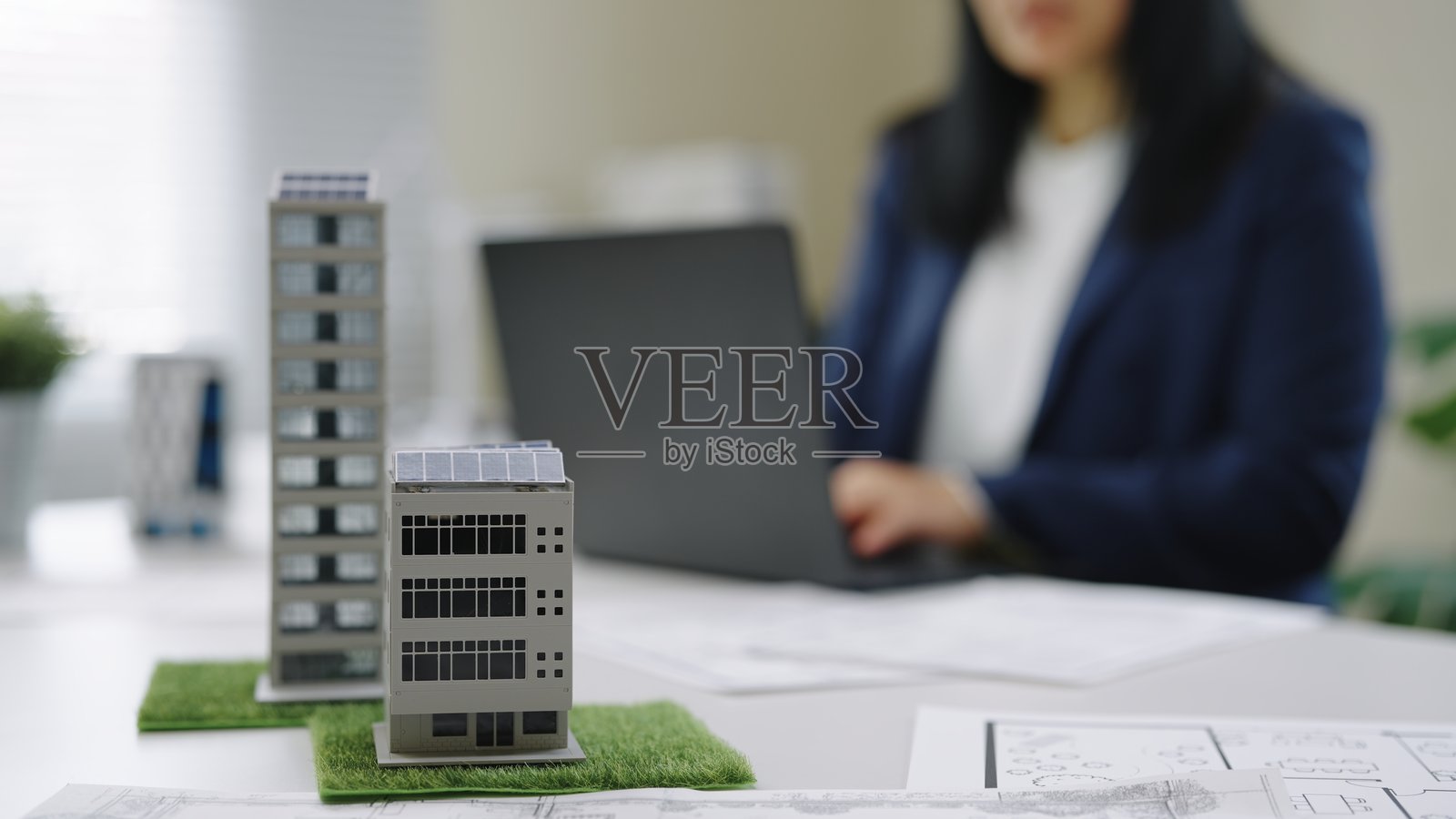 女建筑师在笔记本电脑上工作，旁边是建筑蓝图、办公楼外部和太阳能电池板的 3D 模型，正在进行可持续城市发展规划或设计会议。照片摄影图片