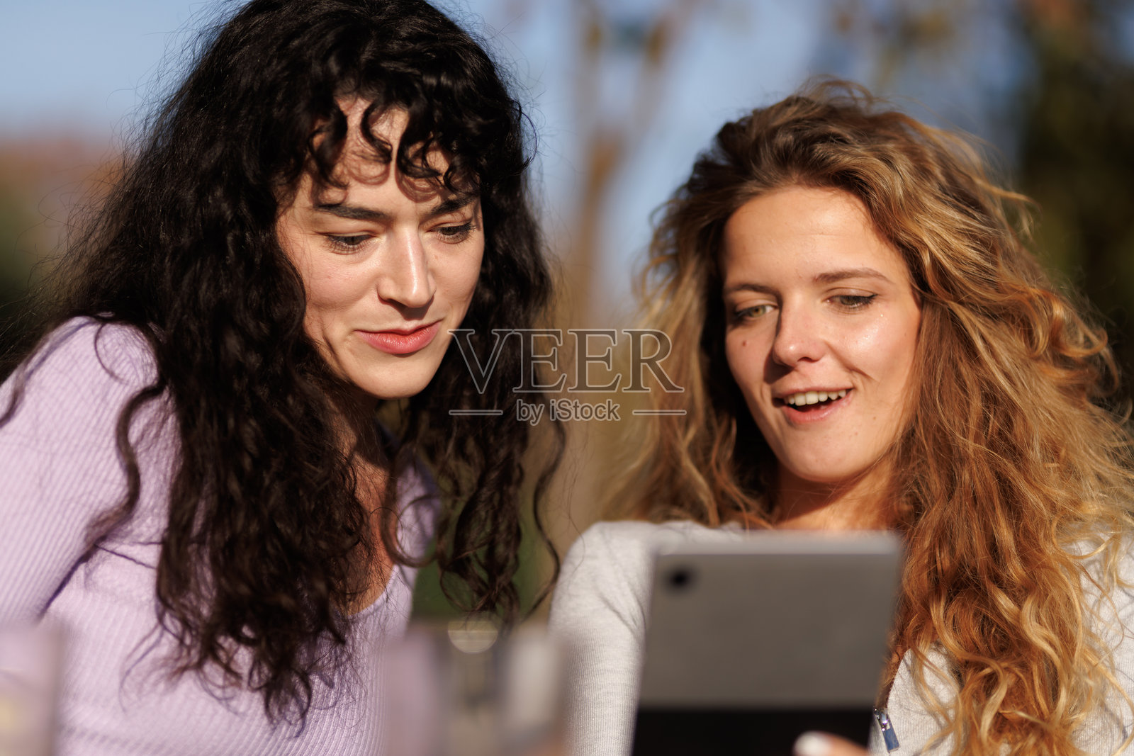 两个朋友在温暖的午后，阳光明媚的公园里，在一次休闲聚会中，一起看着智能手机，享受着户外的时光。照片摄影图片