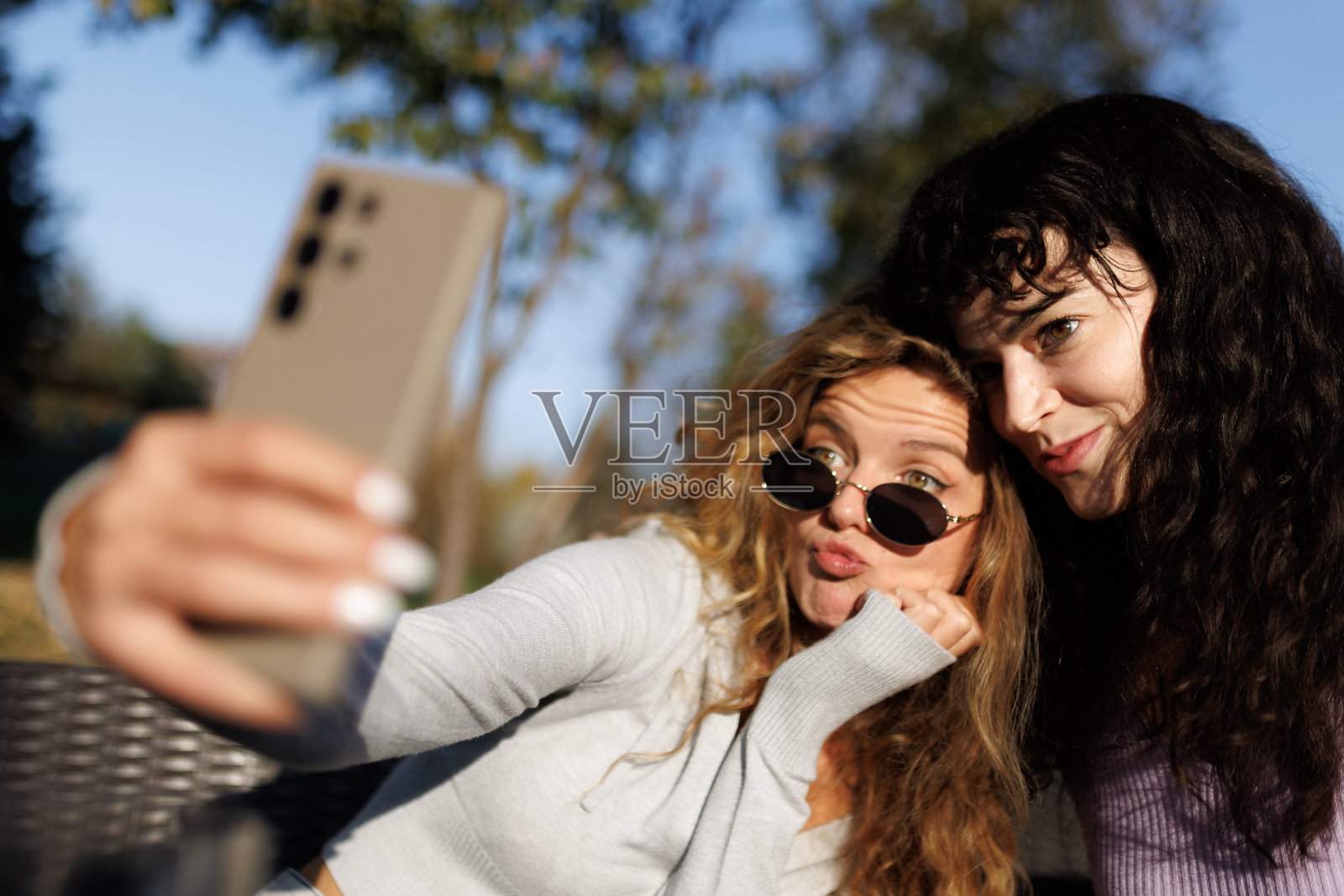 两个朋友在阳光明媚的户外，用智能手机自拍，捕捉公园里充满乐趣的瞬间，背景有树木。照片摄影图片