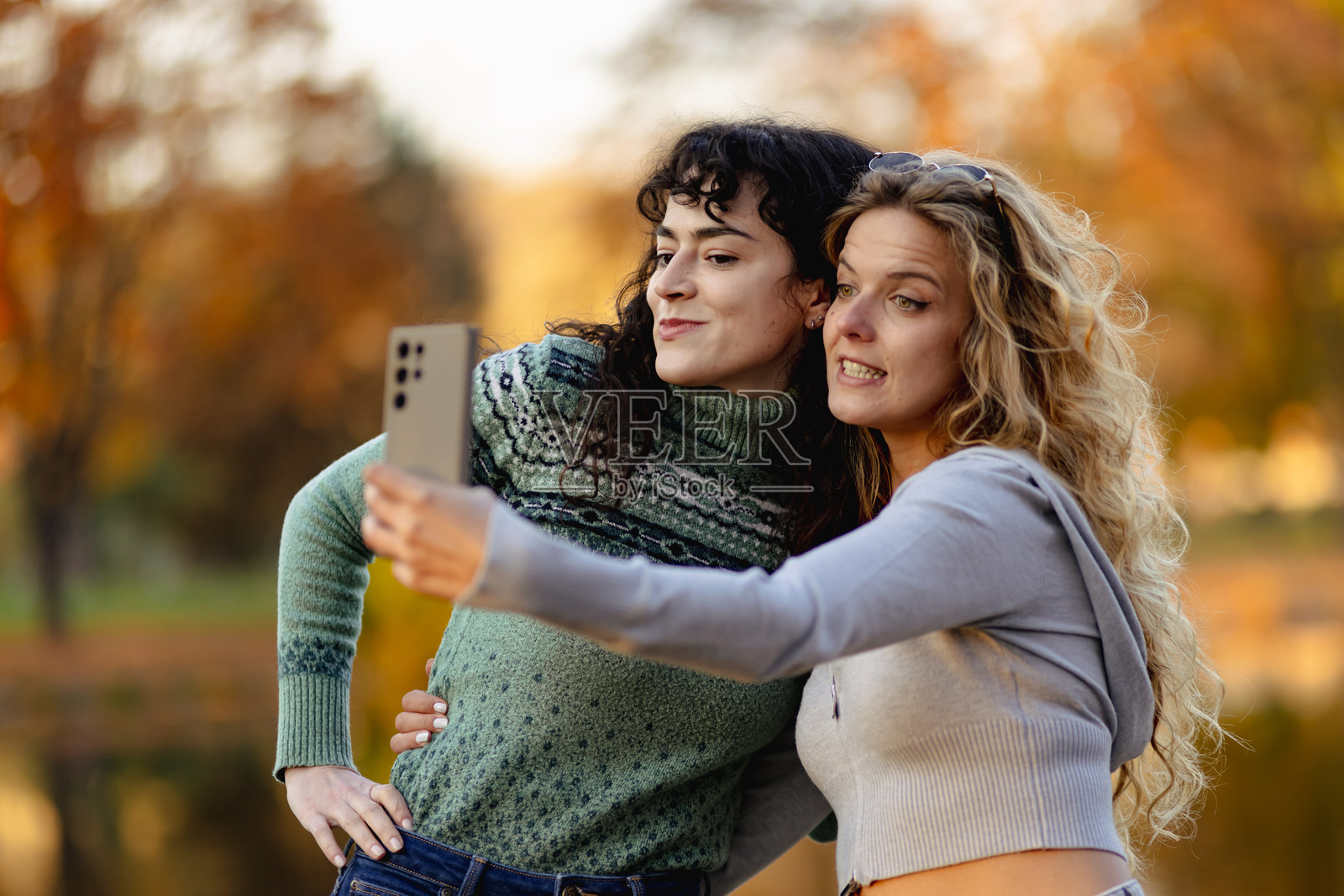 朋友们在公园里享受阳光明媚的一天，在水边自拍，背景是色彩鲜艳的秋叶，营造出欢乐的氛围。照片摄影图片