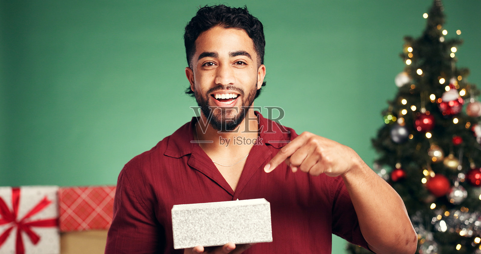 肖像、礼品盒和男人在工作室里，为圣诞节、节日或传统庆祝活动而摆姿势。带着自豪感，男人在圣诞树前，手持礼物，脸上带着快乐的惊喜，背景为绿色，营造出节日气氛。照片摄影图片