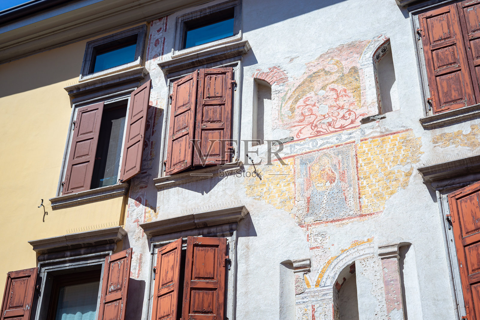 阳光明媚的街道上，一座历史建筑的外墙，配有木制百叶窗和褪色的壁画。照片摄影图片