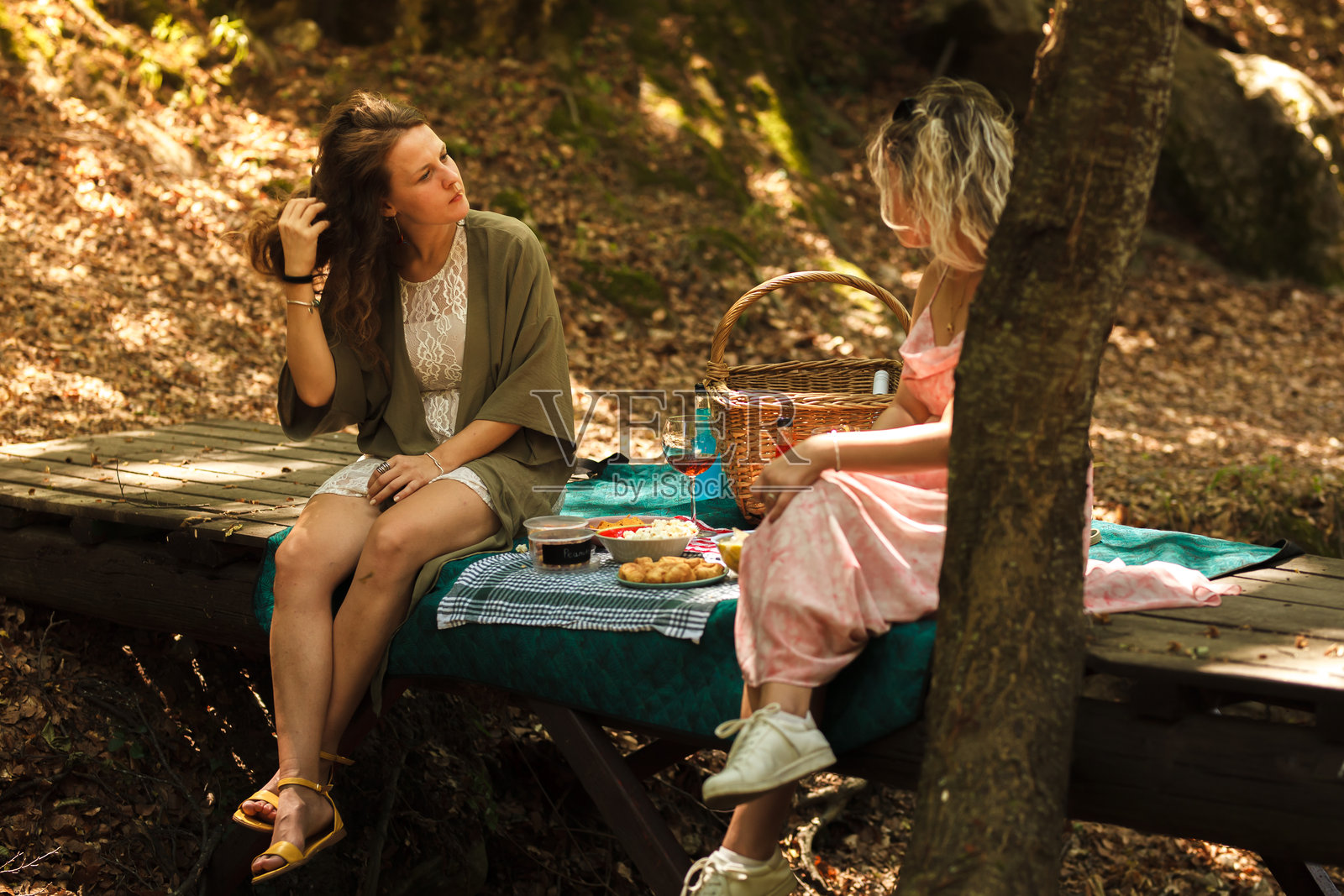 阳光明媚的温暖日子里，朋友们在绿树环绕的木制平台上享受野餐，分享零食并愉快地交谈。照片摄影图片