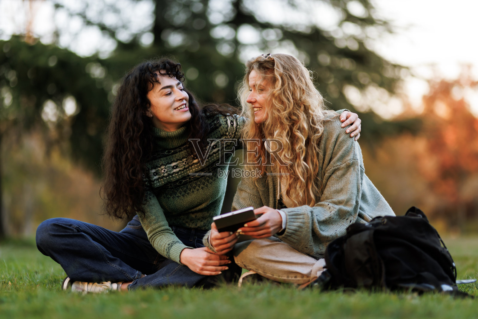 夕阳下，朋友们在公园的草地上放松，共同分享着快乐的时刻，一边欣赏着大自然的美景，一边一起看着平板电脑。照片摄影图片