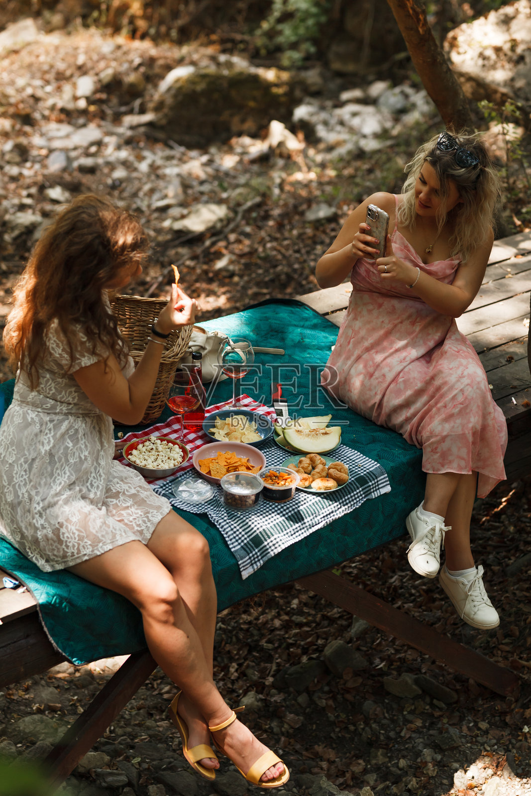 两位好友在宁静的林区享受野餐，在明媚的午后品尝美食并用手机记录美好瞬间。照片摄影图片