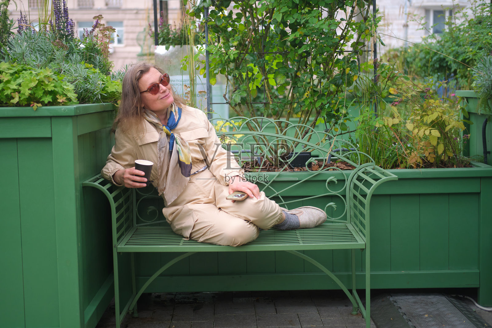 戴着太阳镜的女人坐在绿色长凳上，手里拿着杯子和手机。都市花园和户外咖啡馆概念。中年女性（50岁）照片摄影图片