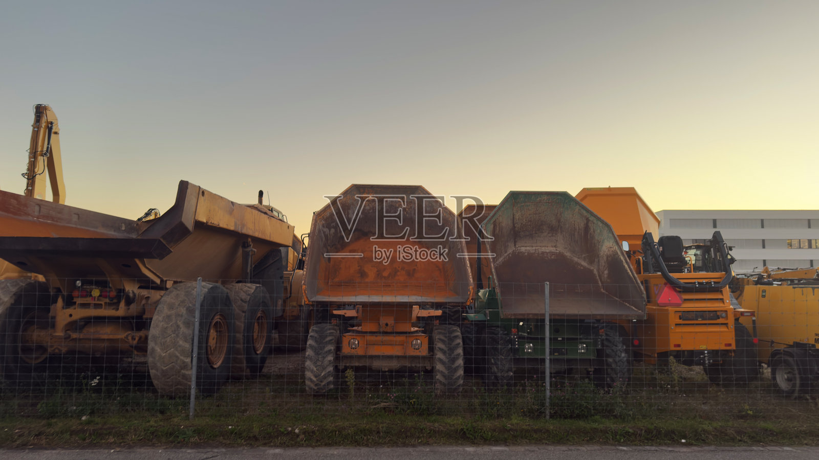 重型建筑机械和建筑材料在日落时分工地上，大型黄色自卸卡车和挖掘机停在金属围栏后面，户外工业设备用于道路工程和土方工程项目照片摄影图片