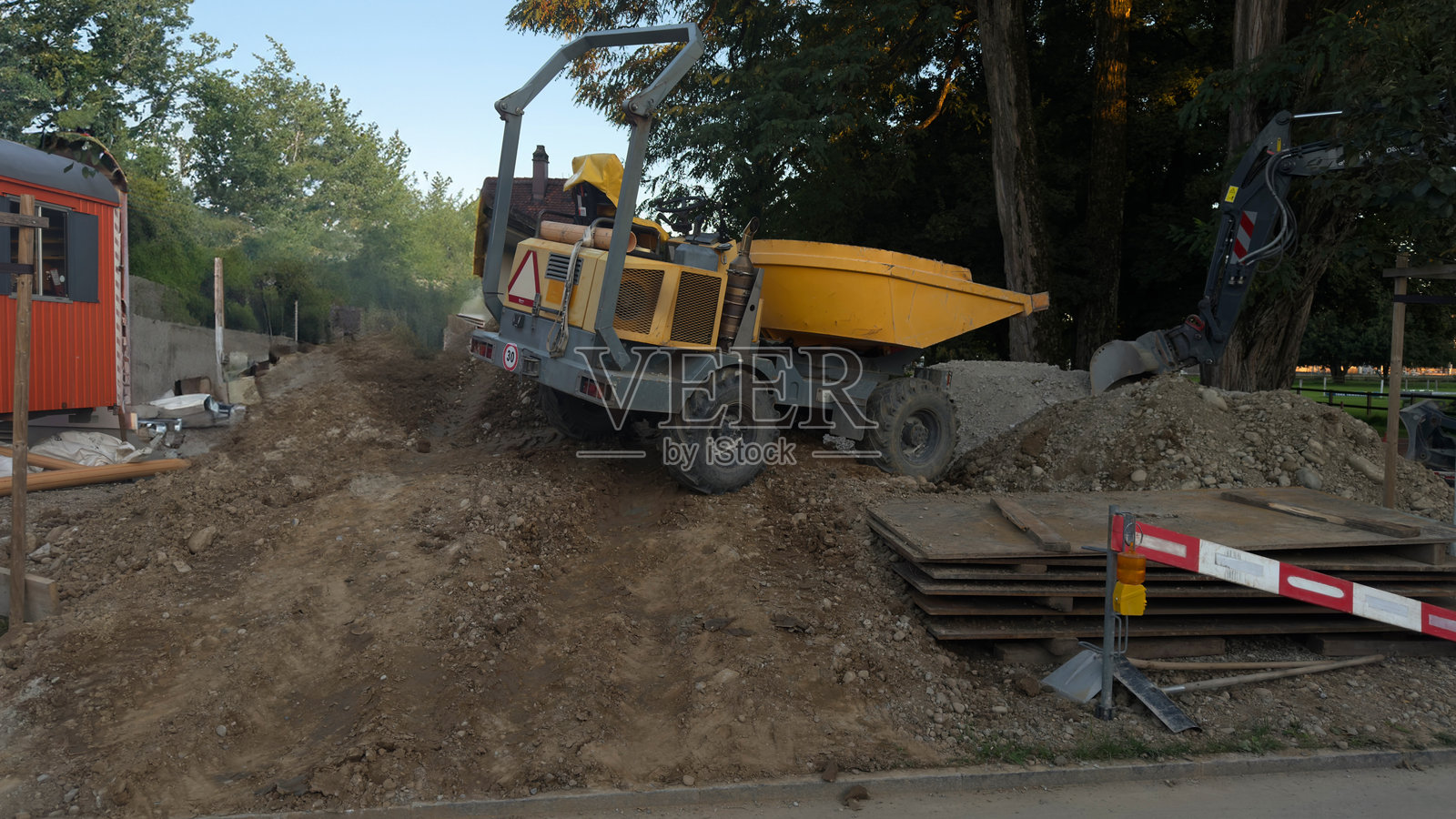 建筑工地，有重型机械和建筑材料，土堆上的黄色自卸卡车，钢板和砾石，背景中有安全屏障和树木，户外道路施工工业设备照片摄影图片