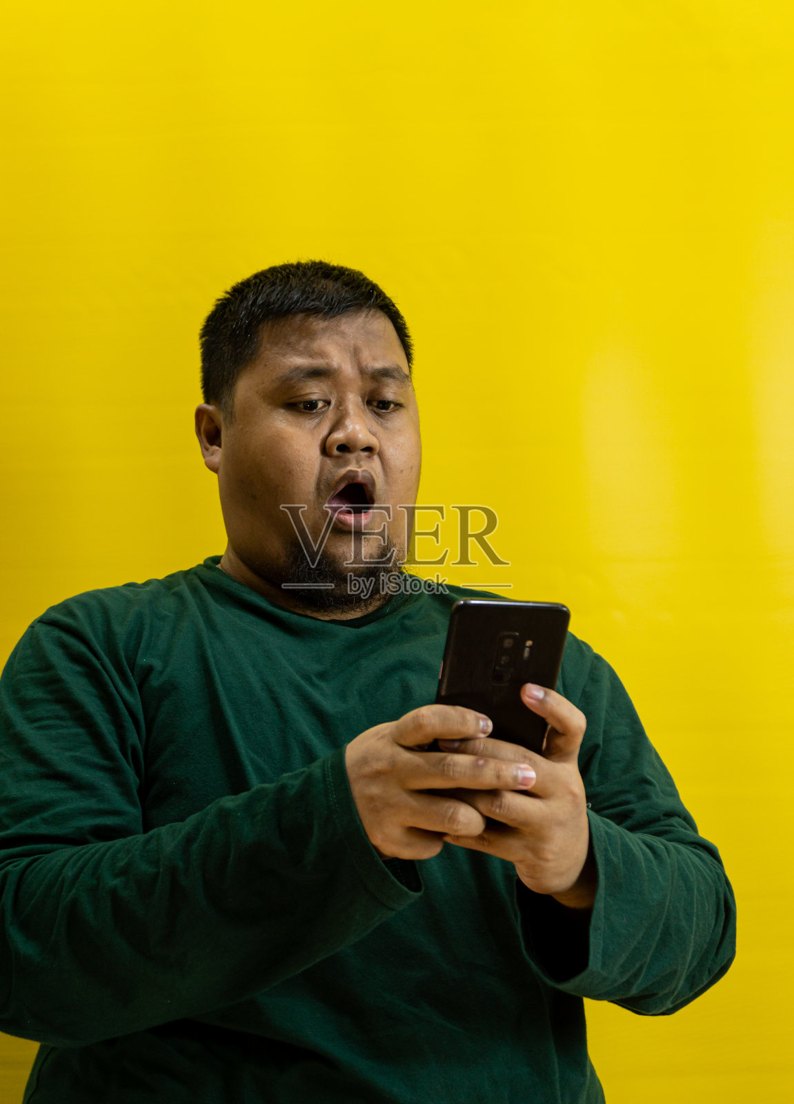 身穿森林绿衬衫的男人，一脸震惊地看着智能手机，孤立在黄色背景上，对消息或意外通知做出惊讶的反应。照片摄影图片