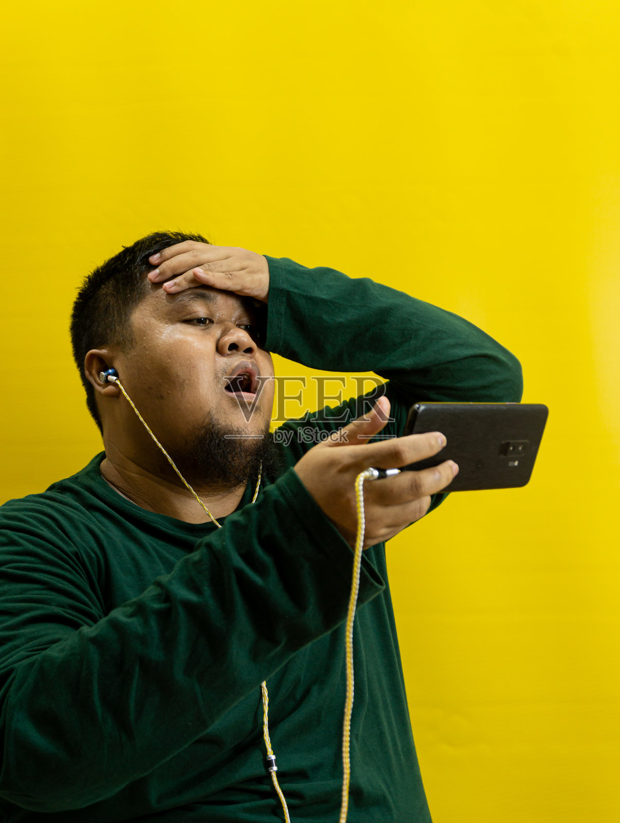穿着绿色衬衫、戴着有线耳机的男人，带着沮丧的表情看着智能手机，一只手扶着额头，孤立在黄色背景上，表现出压力或失望。照片摄影图片