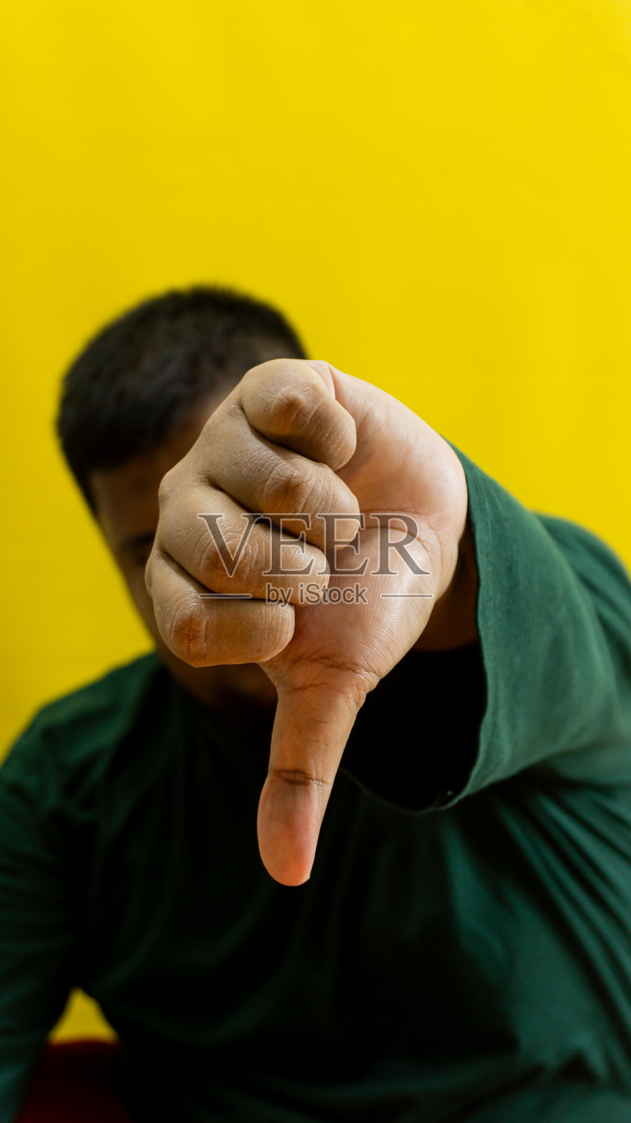 穿着森林绿长袖衬衫的男性模特，做出竖起大拇指向下的手势，背景为黄色，表情清晰地表达不喜欢、不同意或负面意见，独立呈现。照片摄影图片