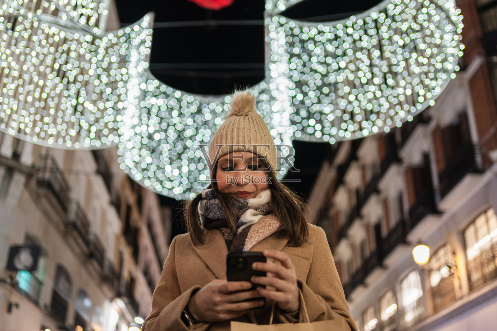 女人在圣诞灯饰下的购物街上使用智能手机照片摄影图片