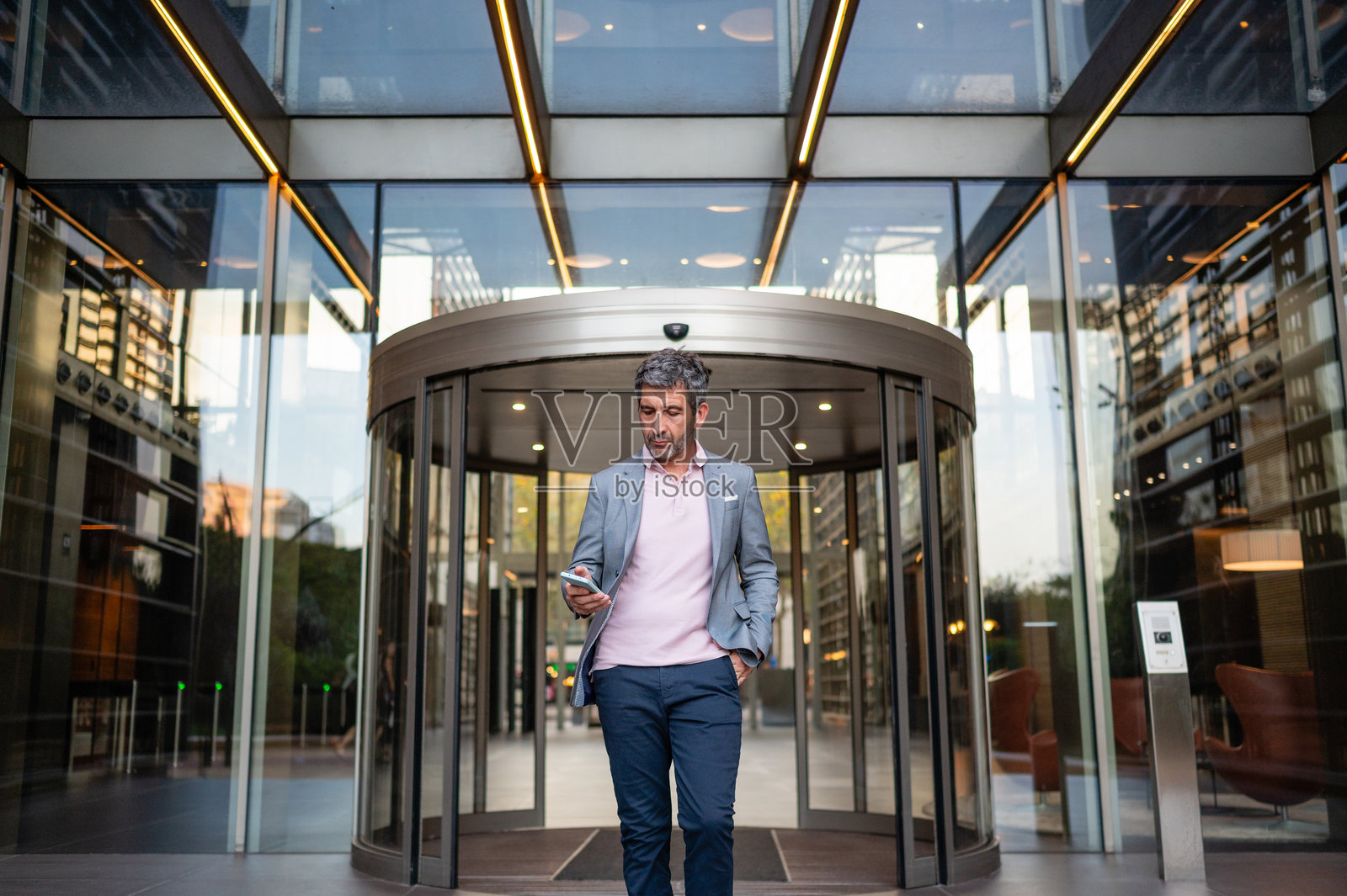 男人从办公楼走出来，一边使用手机。照片摄影图片