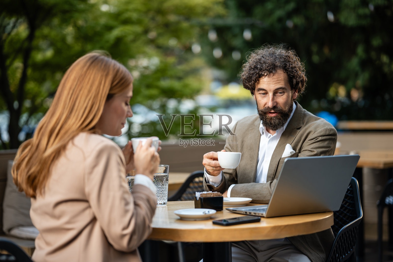 商务人士在户外咖啡馆喝咖啡并讨论笔记本电脑上的工作。照片摄影图片