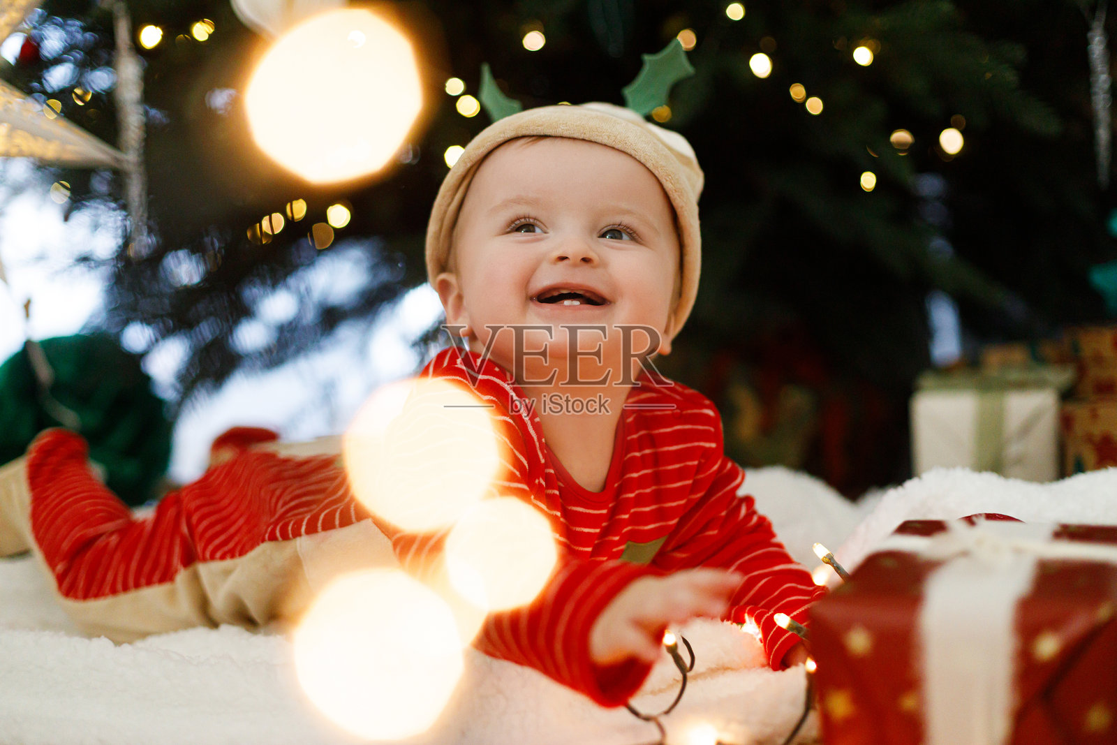 可爱的宝宝穿着圣诞服装，开心地抱着彩灯，躺在装饰时尚的圣诞树和礼物下。圣诞快乐，节日快乐！宝宝的第一个圣诞节。照片摄影图片
