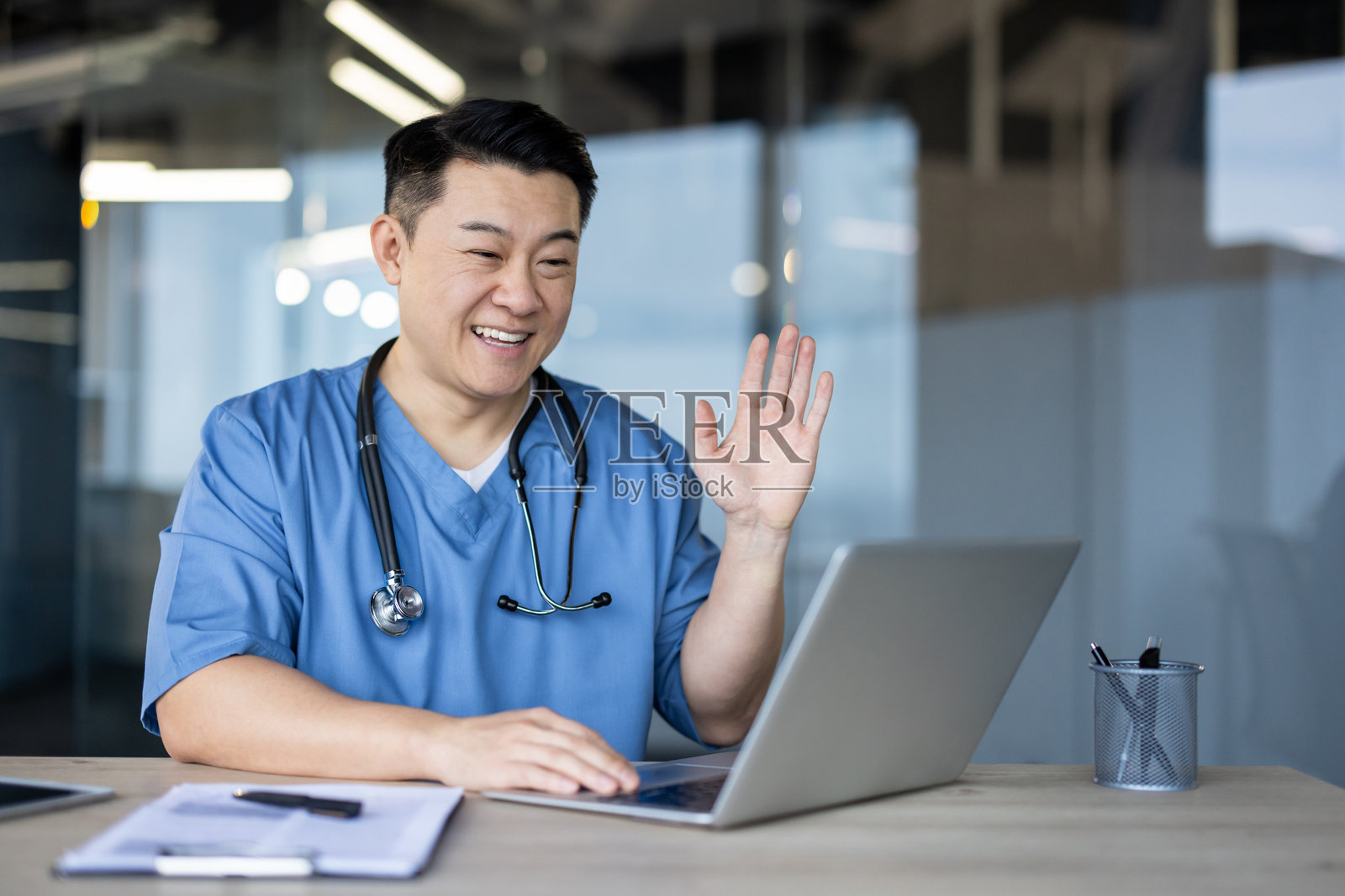 一位亚洲年轻男医生，学生，身穿蓝色制服，坐在医院的桌子旁，对着笔记本电脑摄像头打招呼并交谈。照片摄影图片