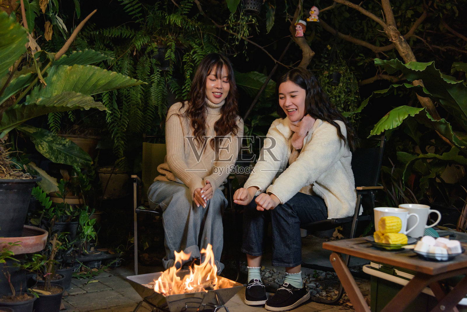 冬日营地户外朋友，森林中的快乐女人，享受夜晚的篝火，朋友们在温暖的冬季假期分享欢声笑语和故事，在茂密的植物中享受热饮和零食的舒适聚会。照片摄影图片