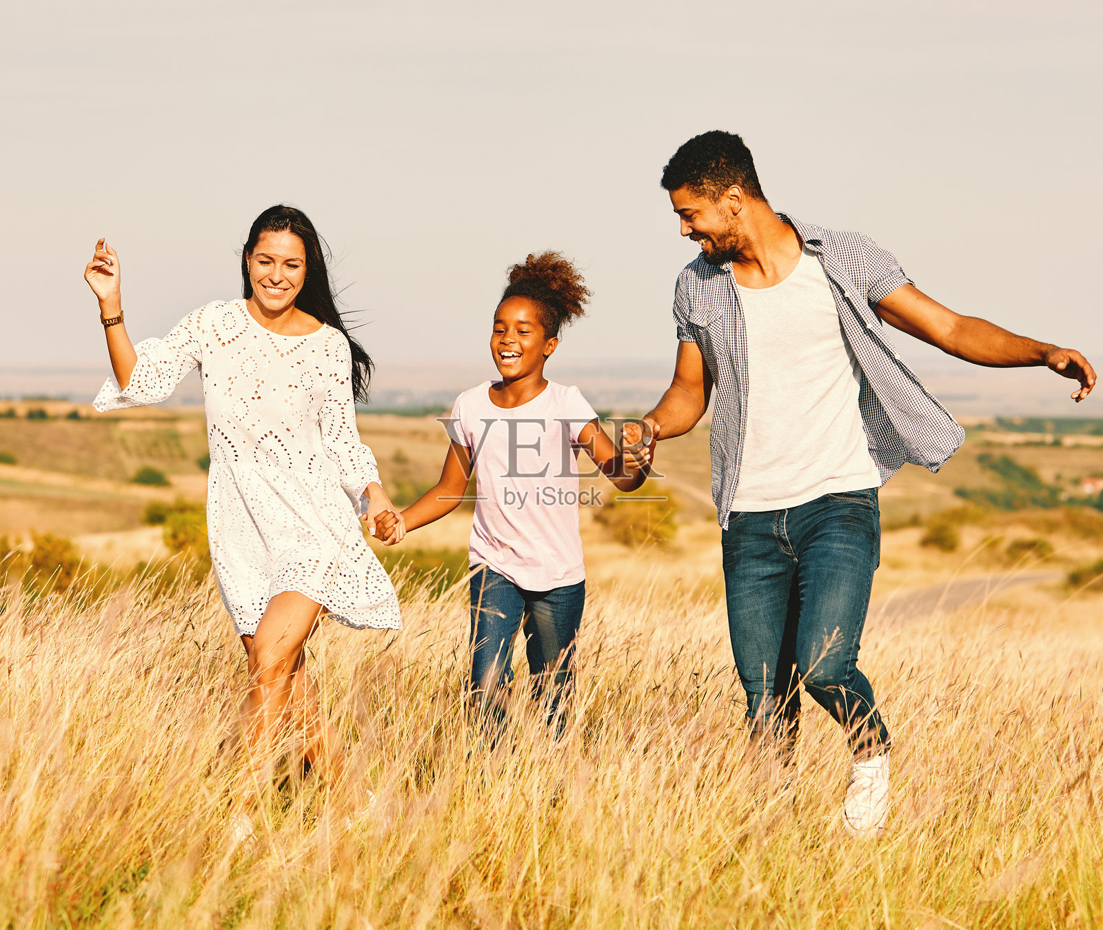 快乐的家庭，父母带着女儿在户外田野里奔跑，享受夏日健康无忧的乐趣，女孩走路时面带笑容，充满活力。照片摄影图片