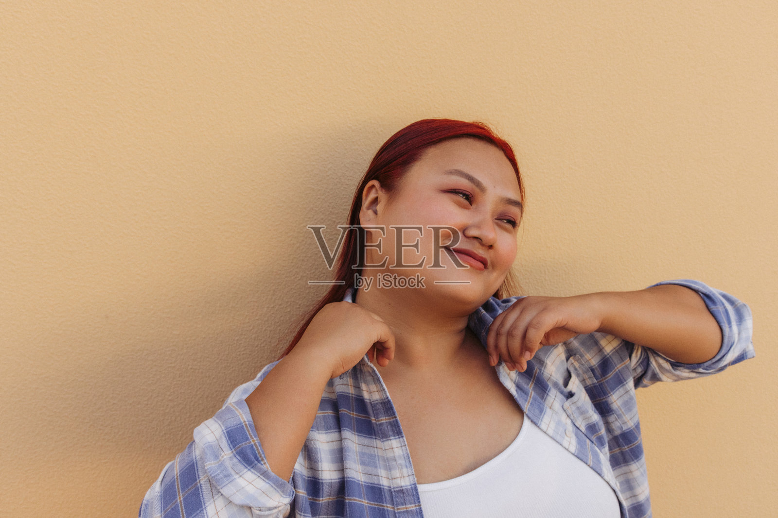 一位红发女子身穿休闲格子衬衫，靠在一个温暖的桃色墙壁旁，温柔地微笑着。照片摄影图片