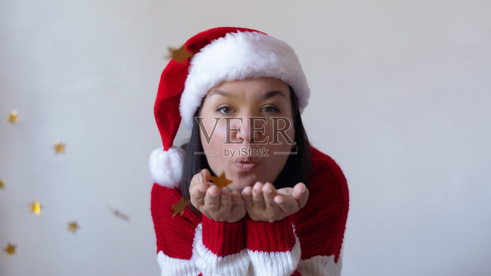戴着圣诞帽、穿着红白条纹毛衣的年轻女子正从手中撒出金色的星星彩纸，在白色背景下营造出欢乐喜庆的节日氛围。照片摄影图片
