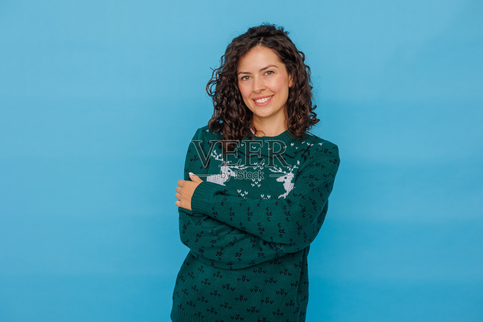 一位年轻女子穿着带有鹿图案的绿色圣诞毛衣，微笑着在蓝色工作室背景前摆姿势。照片摄影图片