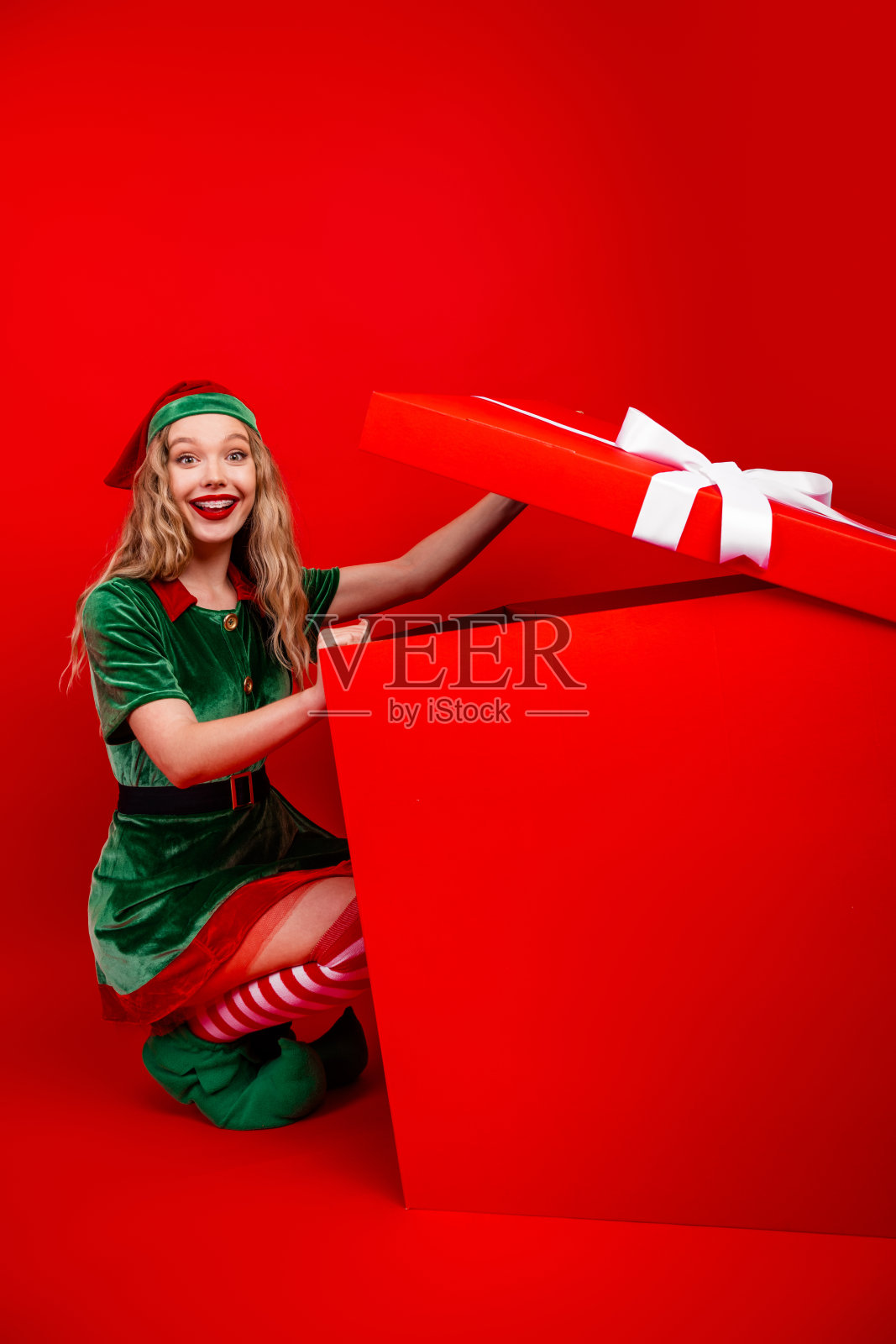 穿着精灵服装的快乐年轻女子在描绘节日喜悦的红色影棚背景前打开一个大礼盒照片摄影图片