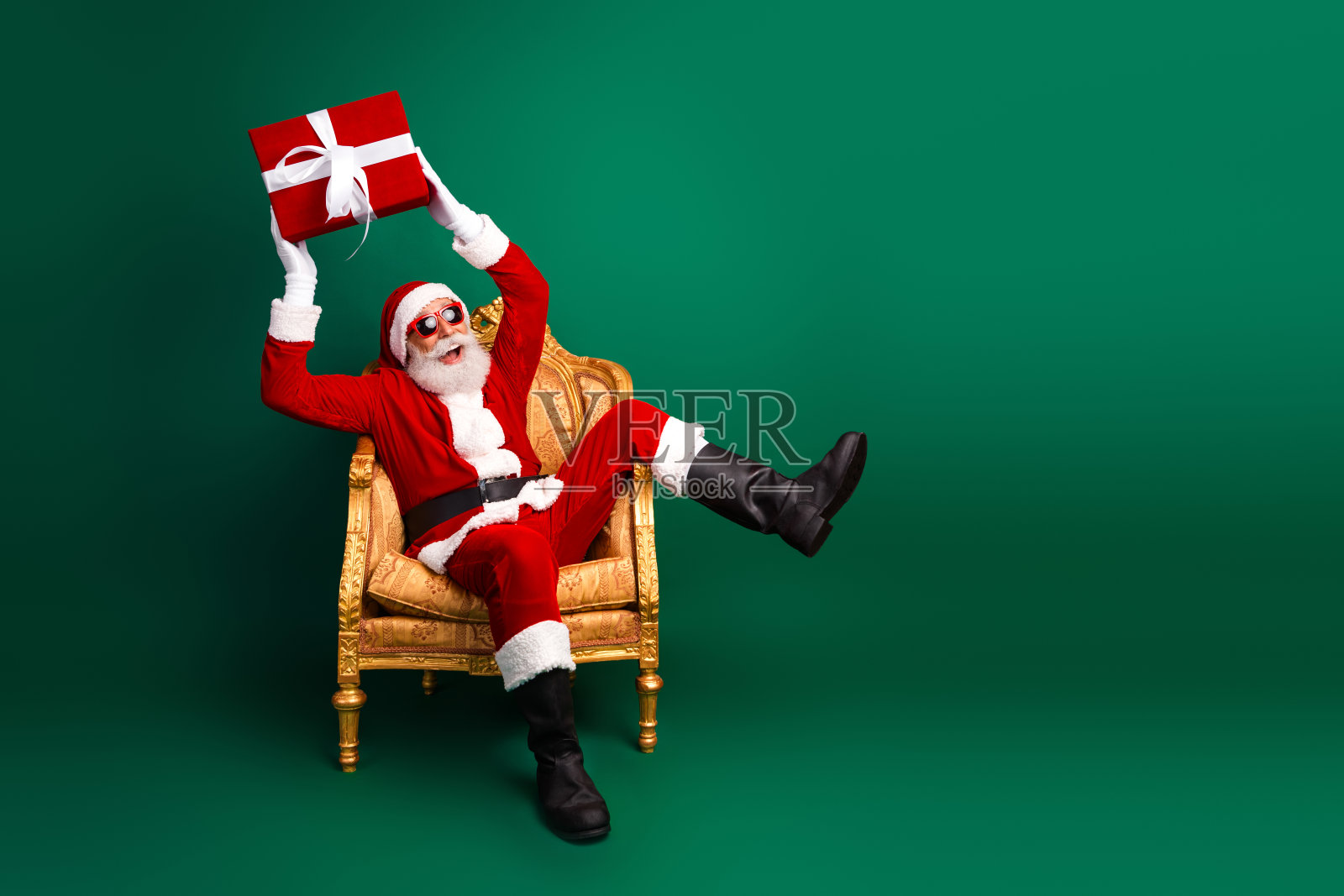 喜庆的圣诞老人坐在一张金色的椅子上，手捧一个包装好的圣诞礼物，背景是节日气氛浓厚的绿色。照片摄影图片