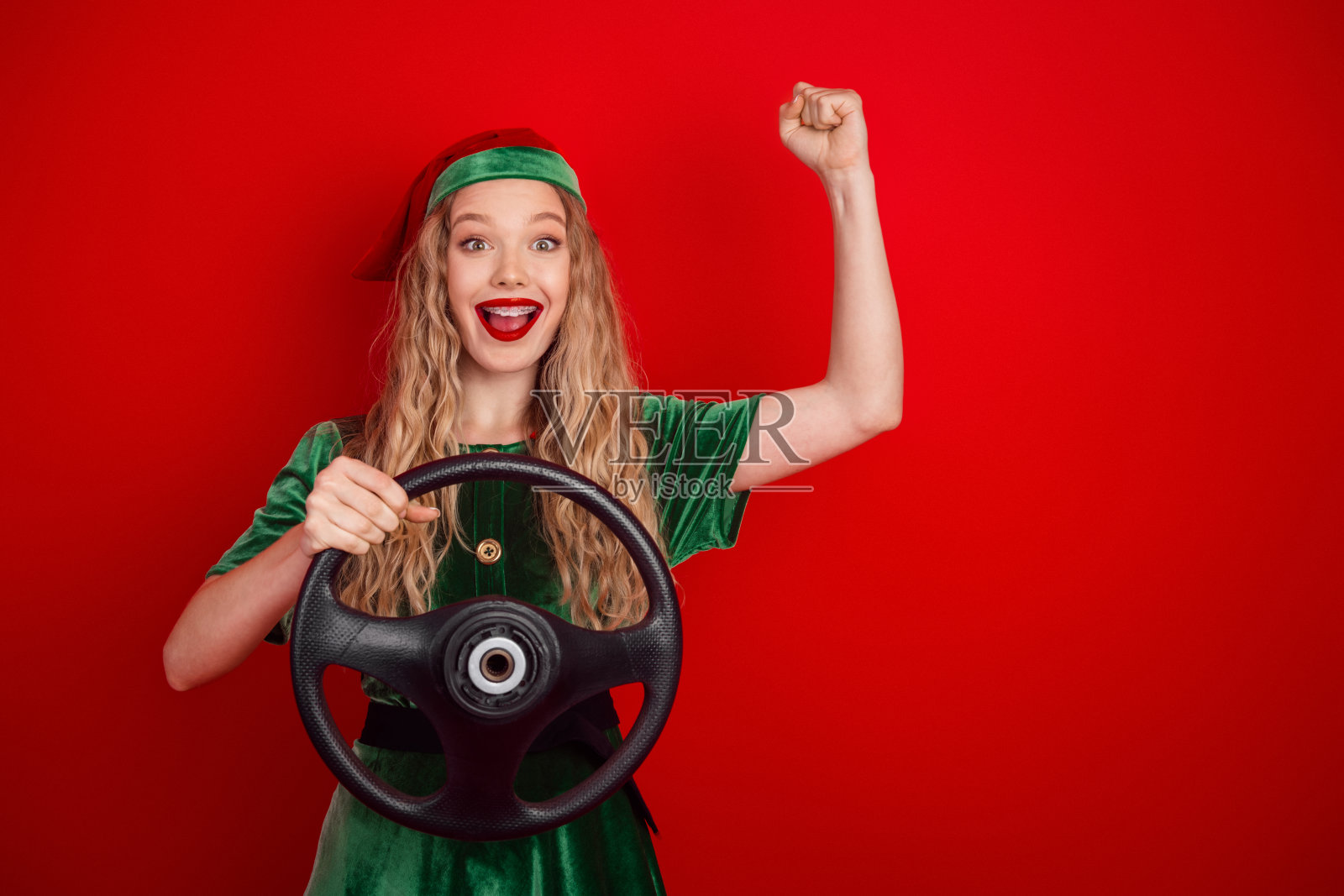 身着圣诞精灵服装的年轻女子兴奋地拿着方向盘，在充满活力的红色背景前庆祝胜利。照片摄影图片