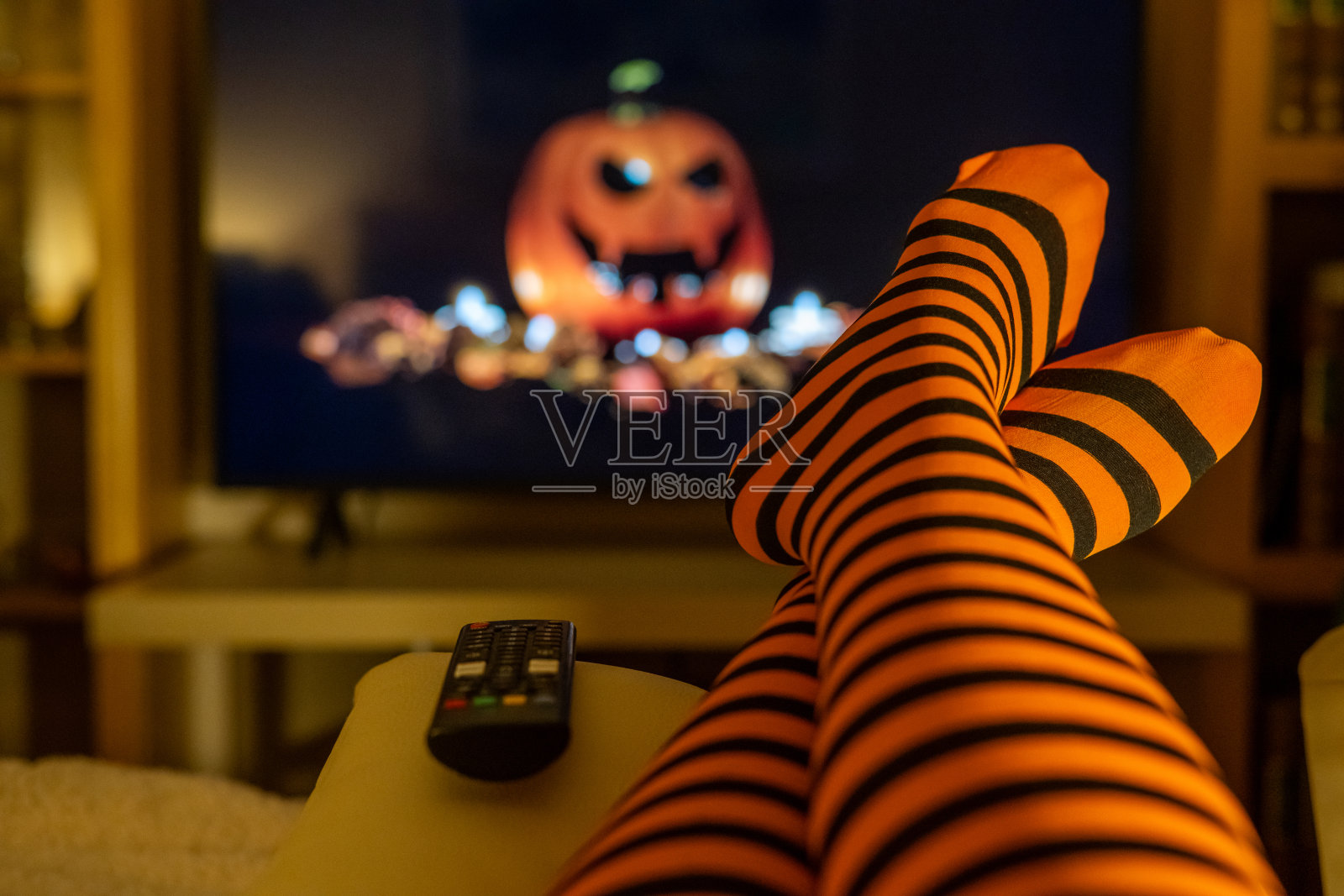 万圣节主题的流媒体内容，在舒适的居家环境中播放，画面中还有橙色条纹的紧身裤，模糊的电视屏幕，营造出一种室内的生活方式和温暖的季节氛围。照片摄影图片