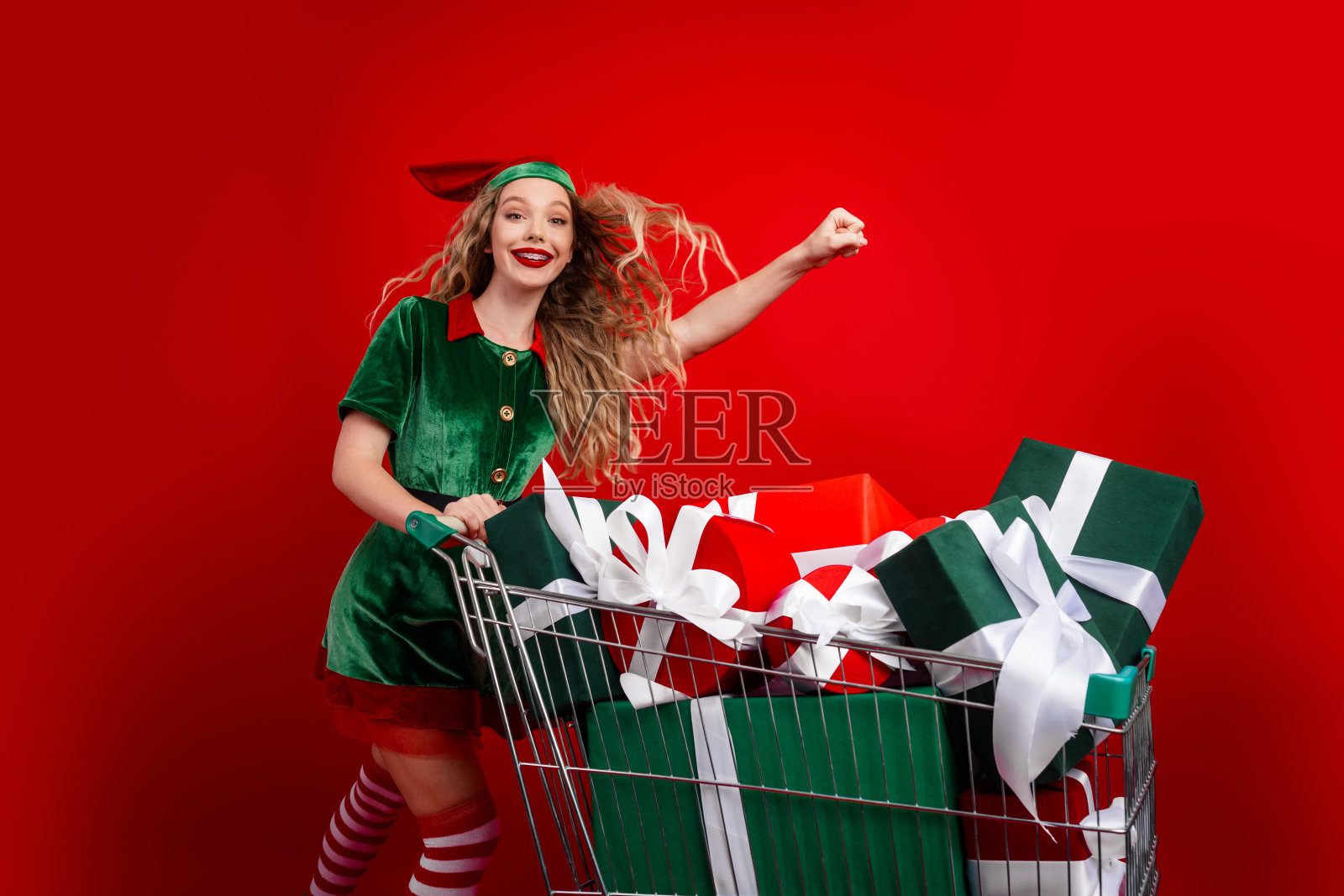 年轻女子打扮成快乐的精灵，推着装满圣诞礼物的购物车，背景是鲜艳的红色。照片摄影图片